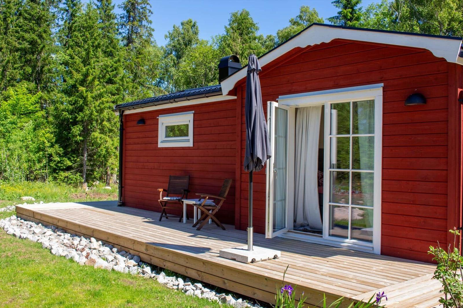Rotes Holzhaus mit Terrasse, Sitzgelegenheit und Gartentisch. Hintergrund: Wald.
