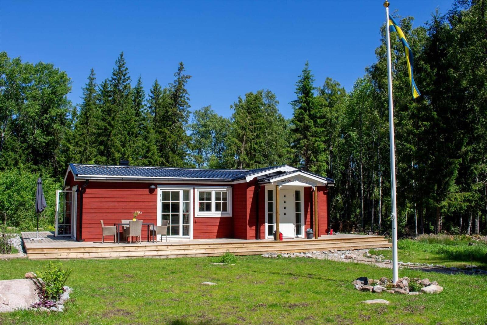 Rotes Holzhaus mit Terrasse, Garten und Flagge vor Wald.