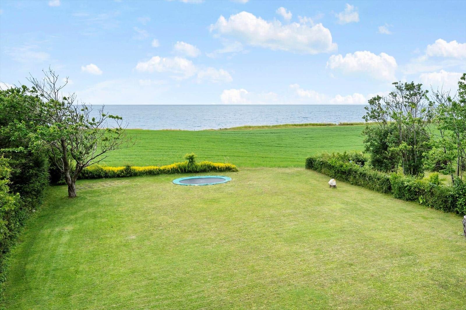 Großer Garten mit Trampolin und Meerblick. Grünfläche, Bäume und Himmel.