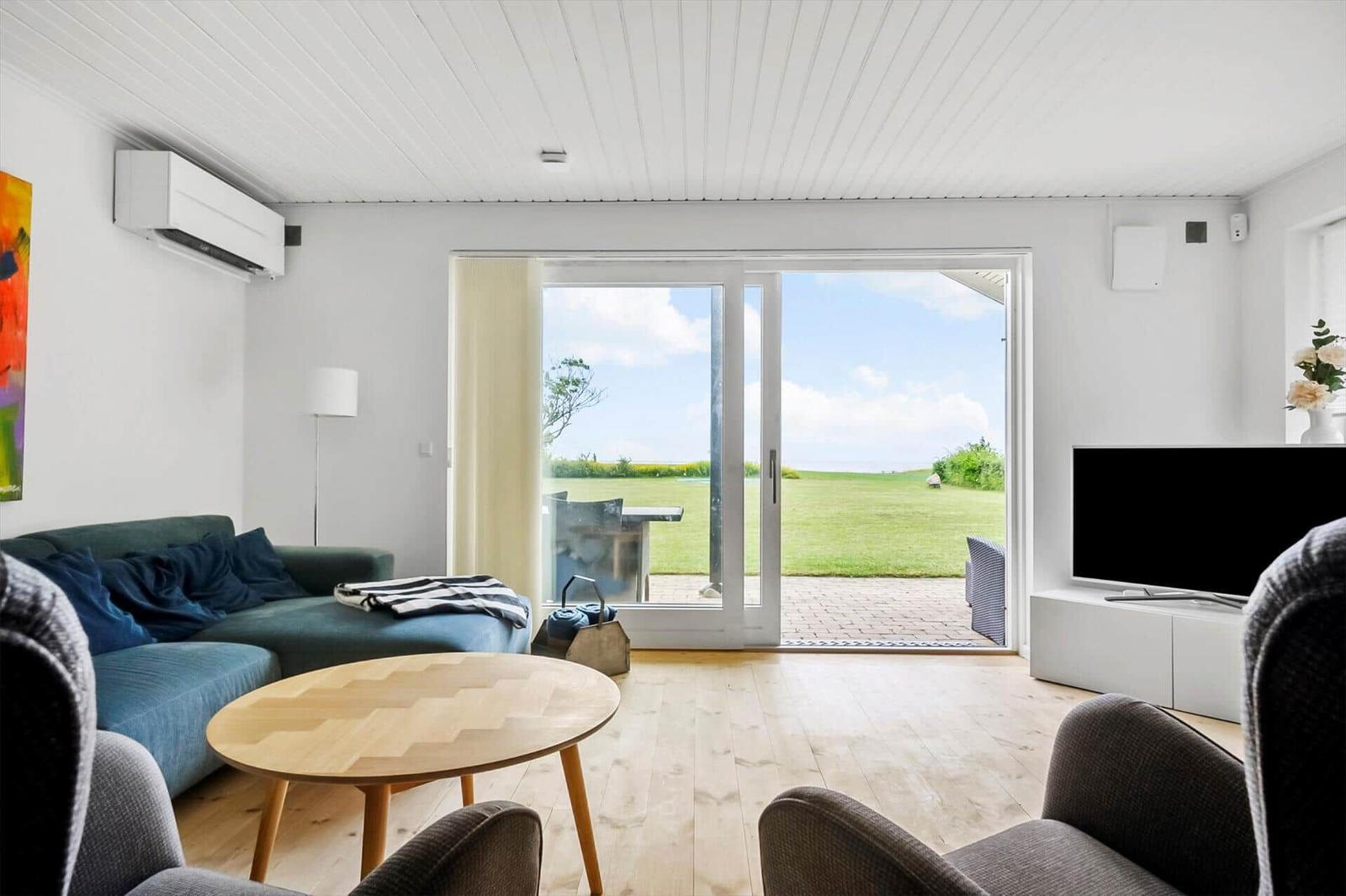 Wohnzimmer mit Sofa, Tisch und TV. Durchgehende Glastür führt auf eine Terrasse mit Blick auf Grasland.
