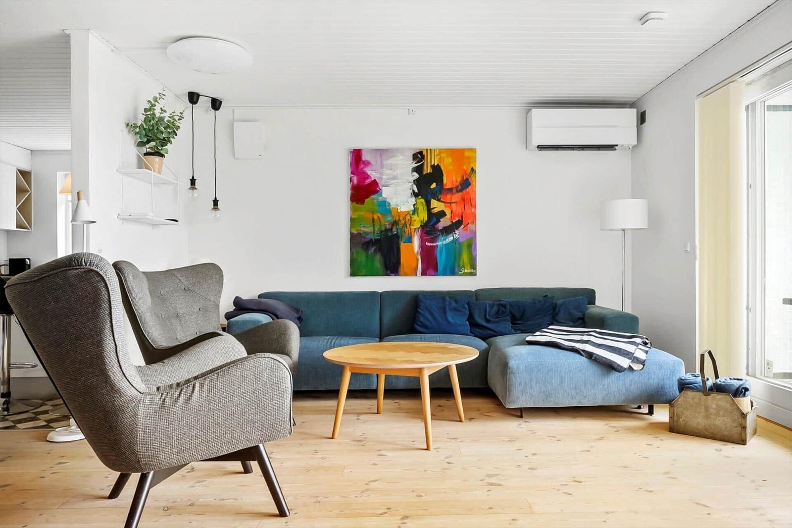 Ein Wohnzimmer mit blauem Sofa, grauer Sessel und Holztisch. An der Wand hängt ein abstraktes Gemälde.