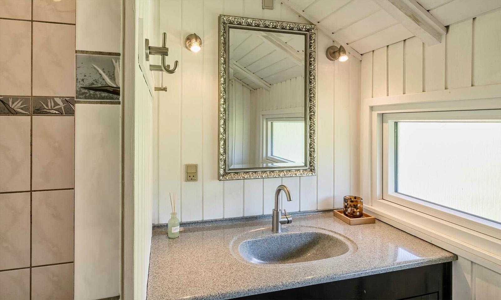 Badezimmer mit Waschbecken, Spiegel und Fenster. Wandverkleidung und Dachbalken sichtbar.
