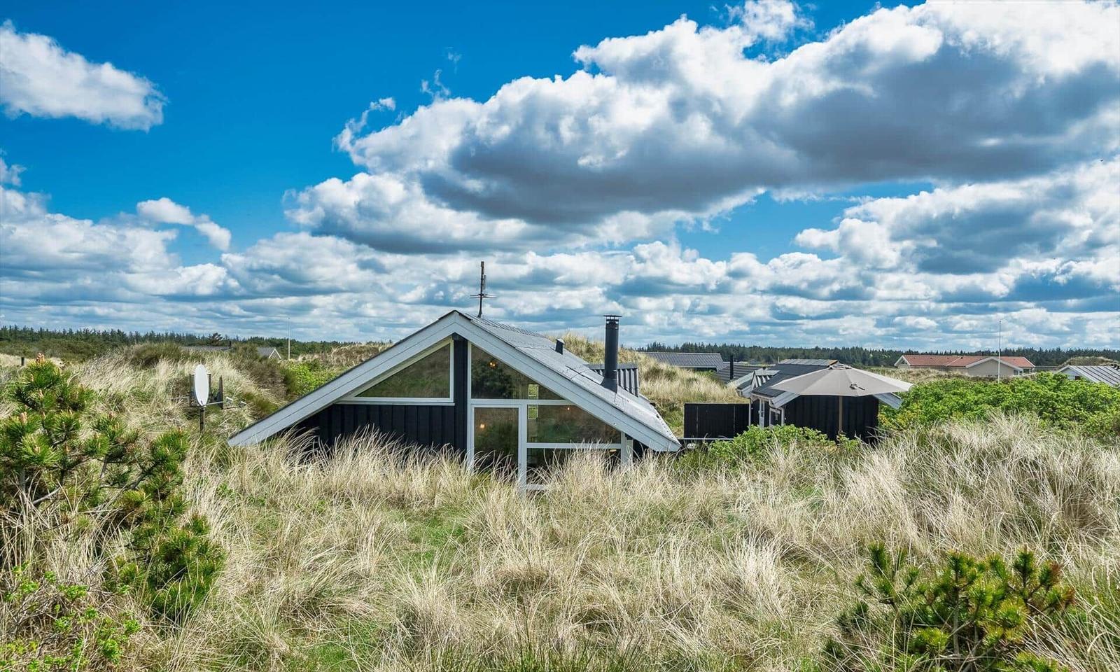 Moderne Hütte mit Dachgaube, umgeben von Gras und Bäumen unter blauem Himmel mit Wolken.