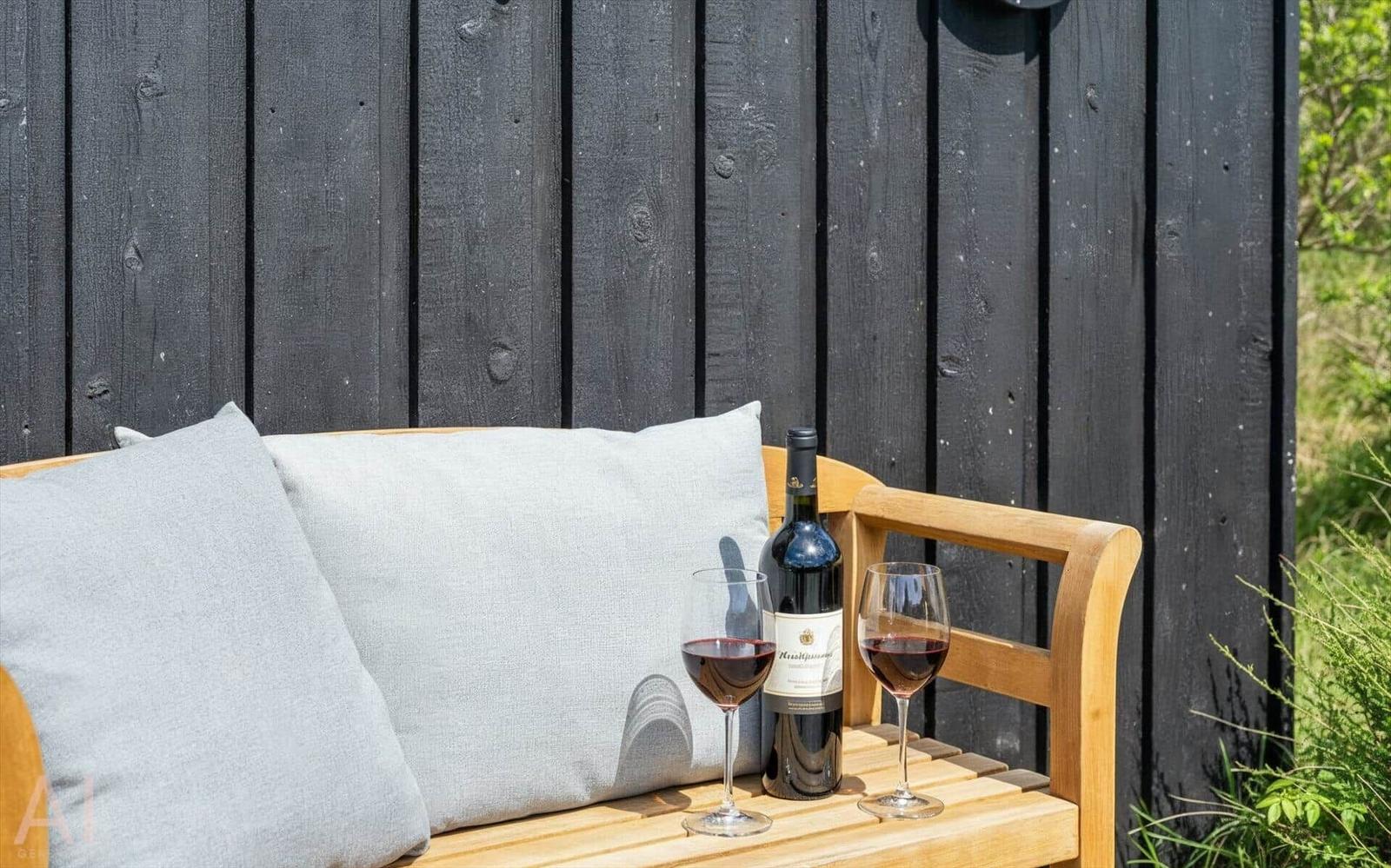 Holzbank mit Kissen, Weinflasche und zwei Gläser auf einer Terrasse.