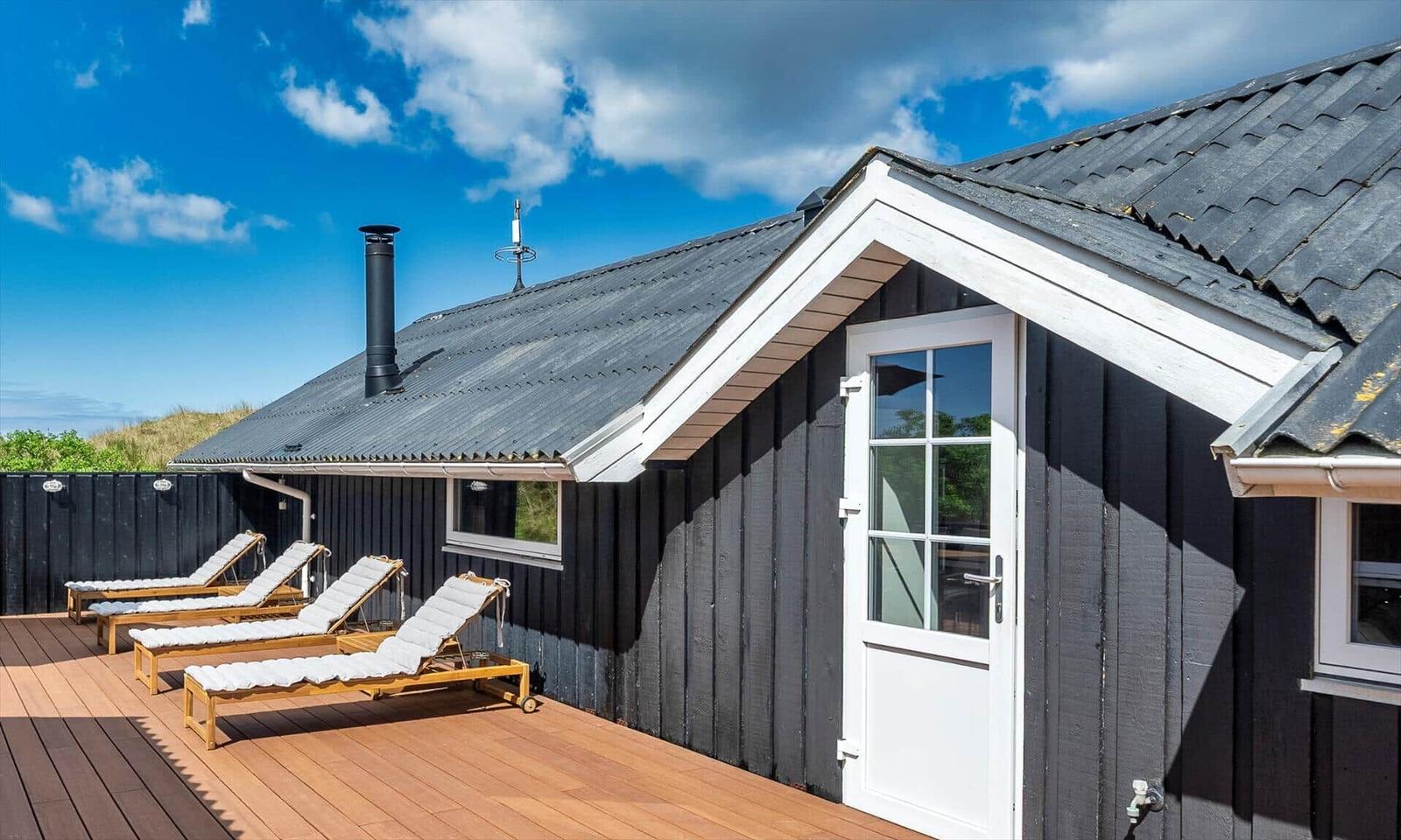 Schwarzes Holzhaus mit Terrasse und Liegestühlen unter blauem Himmel.