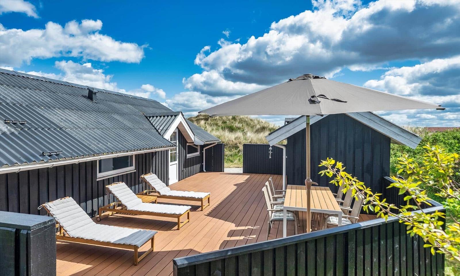 Terrasse mit Liegestühlen, Tisch und Sonnenschirm vor einem Haus mit Holzverkleidung.