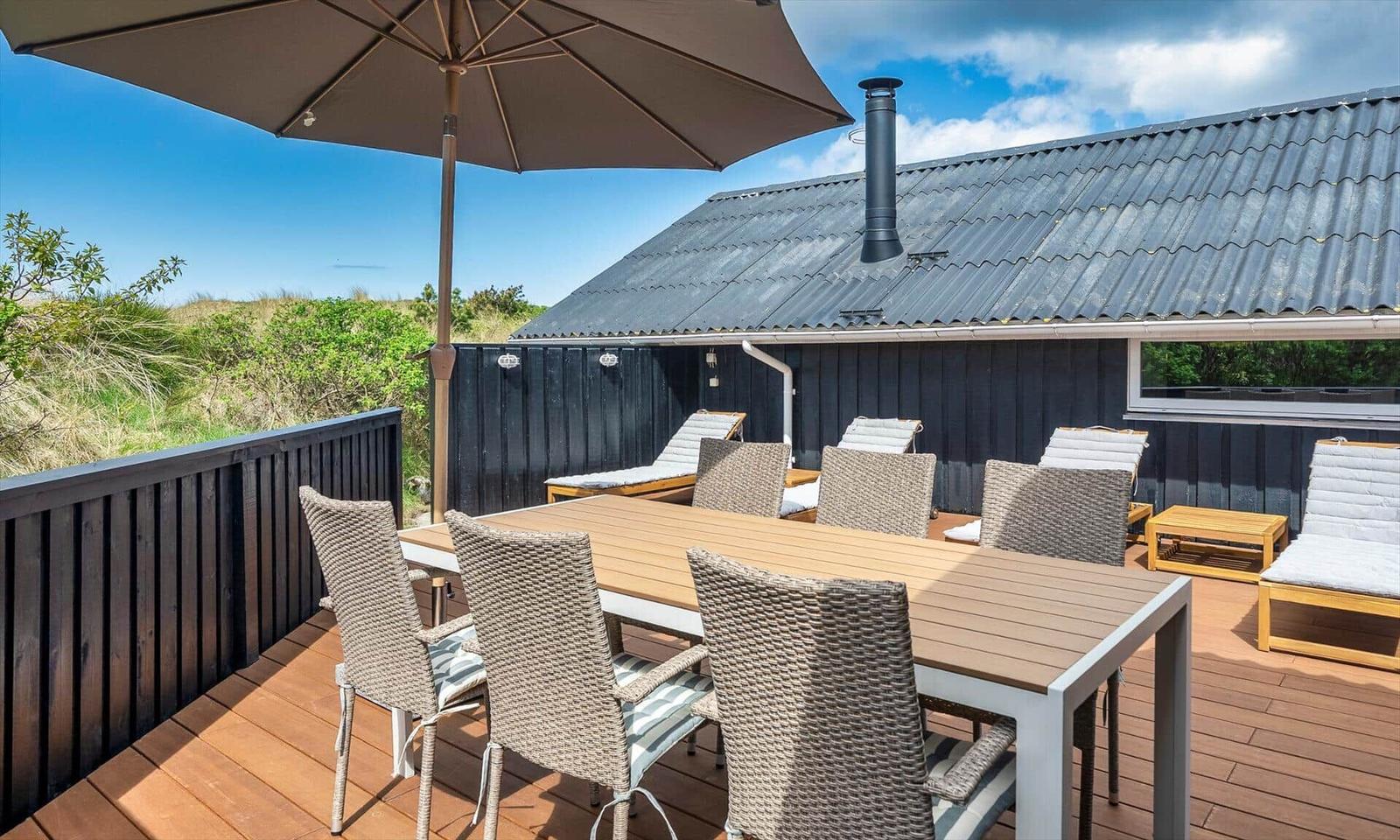 Terrasse mit Tisch, Stühlen und Sonnenschirm. Hintergrund: Haus mit dunklem Holz und Wellblechdach.