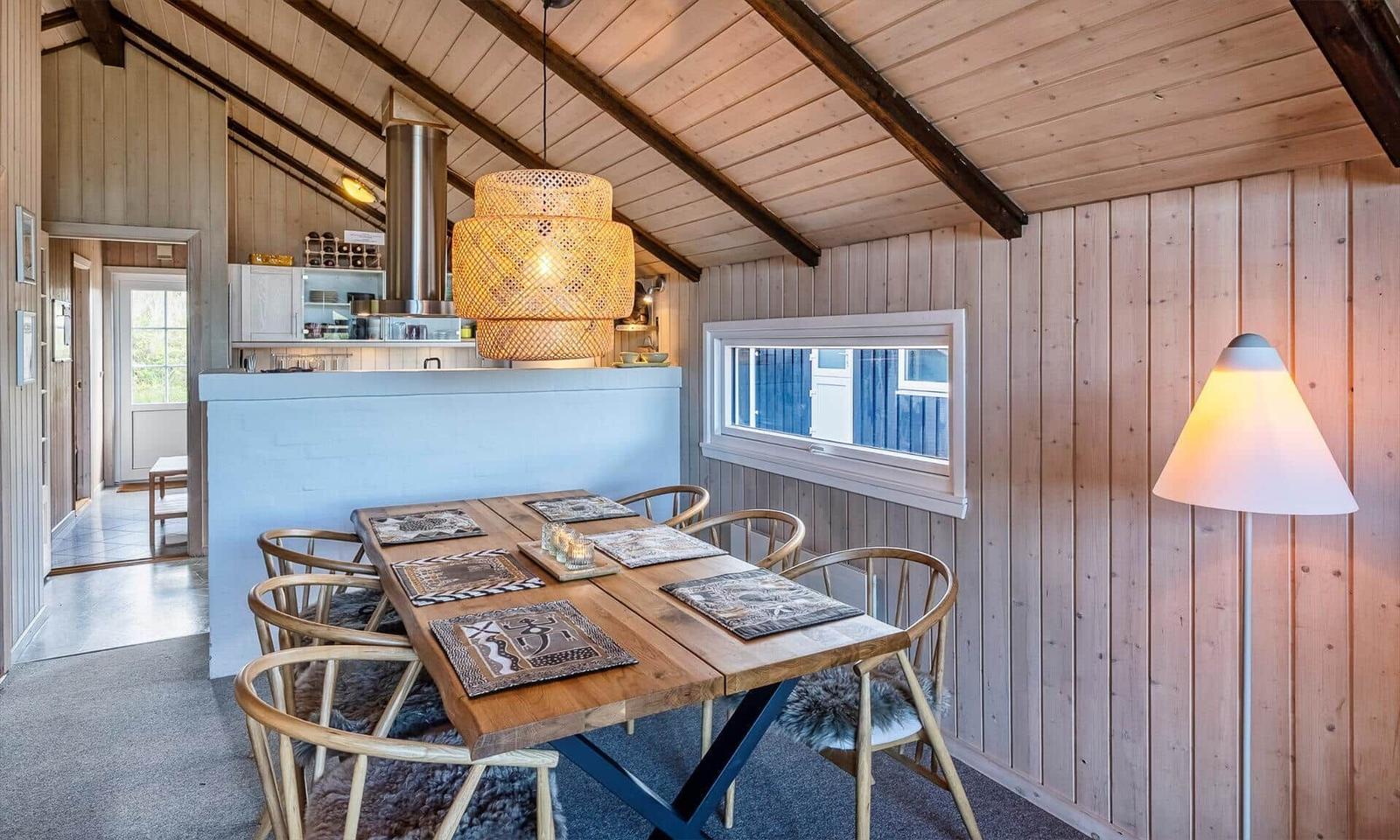 Essbereich mit Holztisch, Stühlen und Lampe. Küche im Hintergrund. Holzwände und Dachbalken.