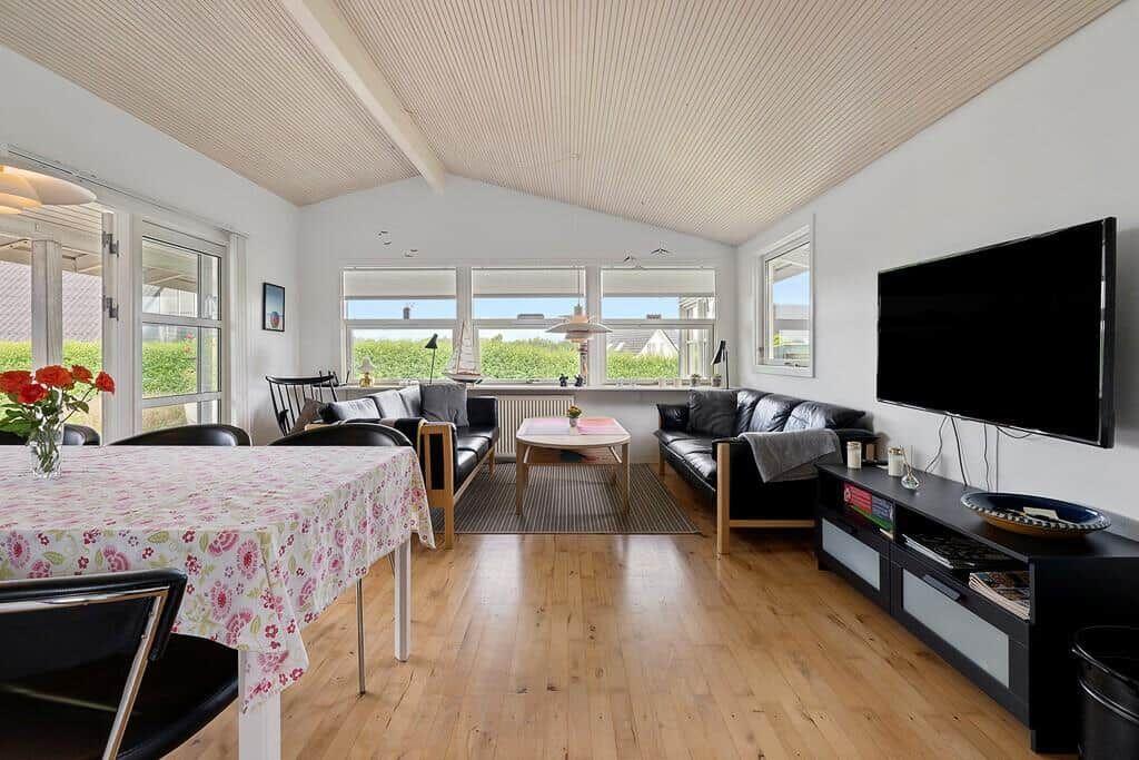 Wohn- und Essbereich mit Holzboden, schwarzen Sofas und TV. Fenster bieten Blick auf Grün.