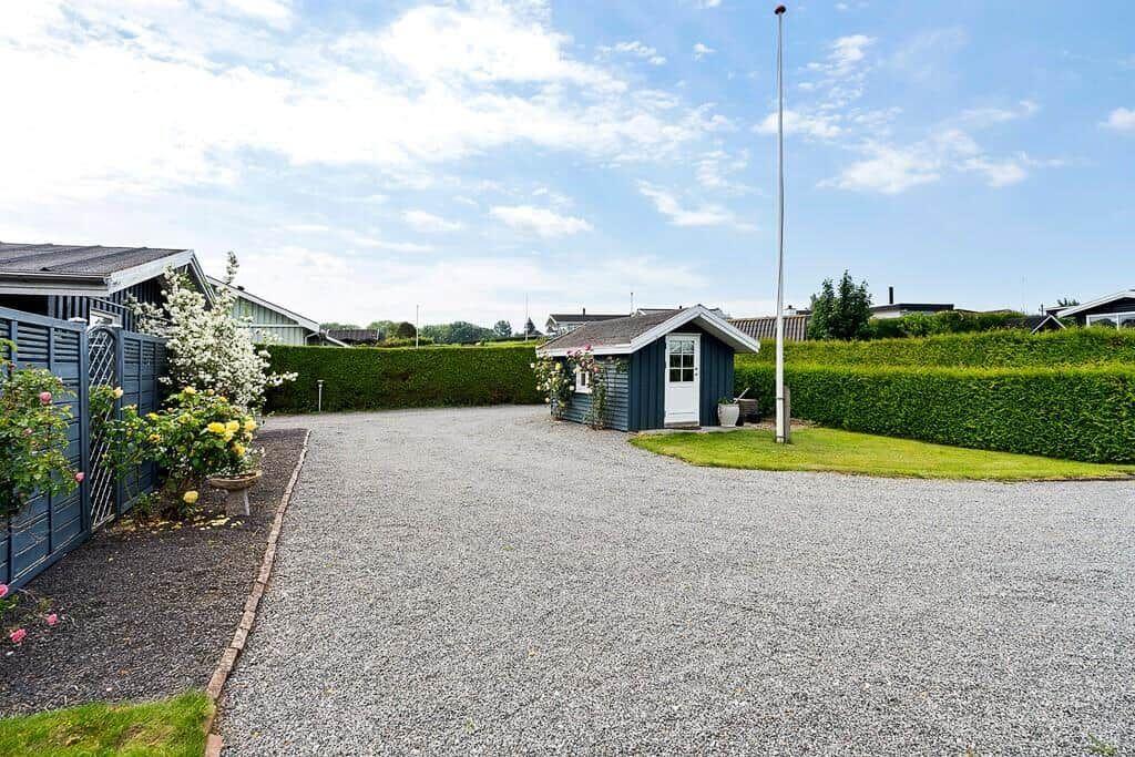 Ein kleines Haus mit weißen Türen steht neben einem Kiesweg. Grüner Zaun und Blumen sind sichtbar.