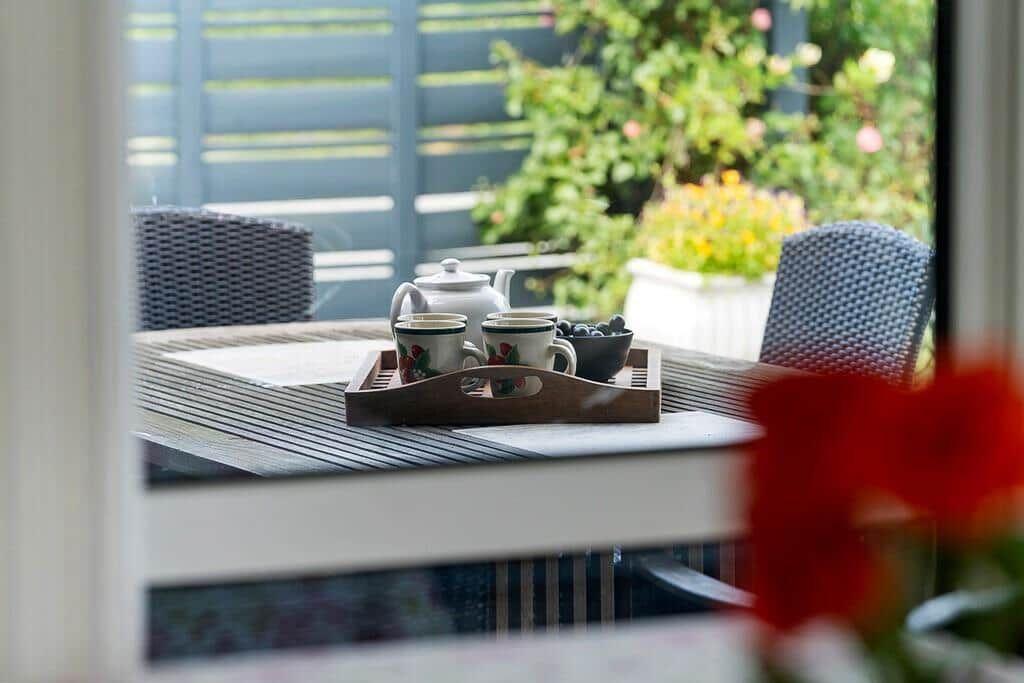 Ein Tisch mit Teekanne und Tassen auf einer Terrasse mit Stühlen und Pflanzen.