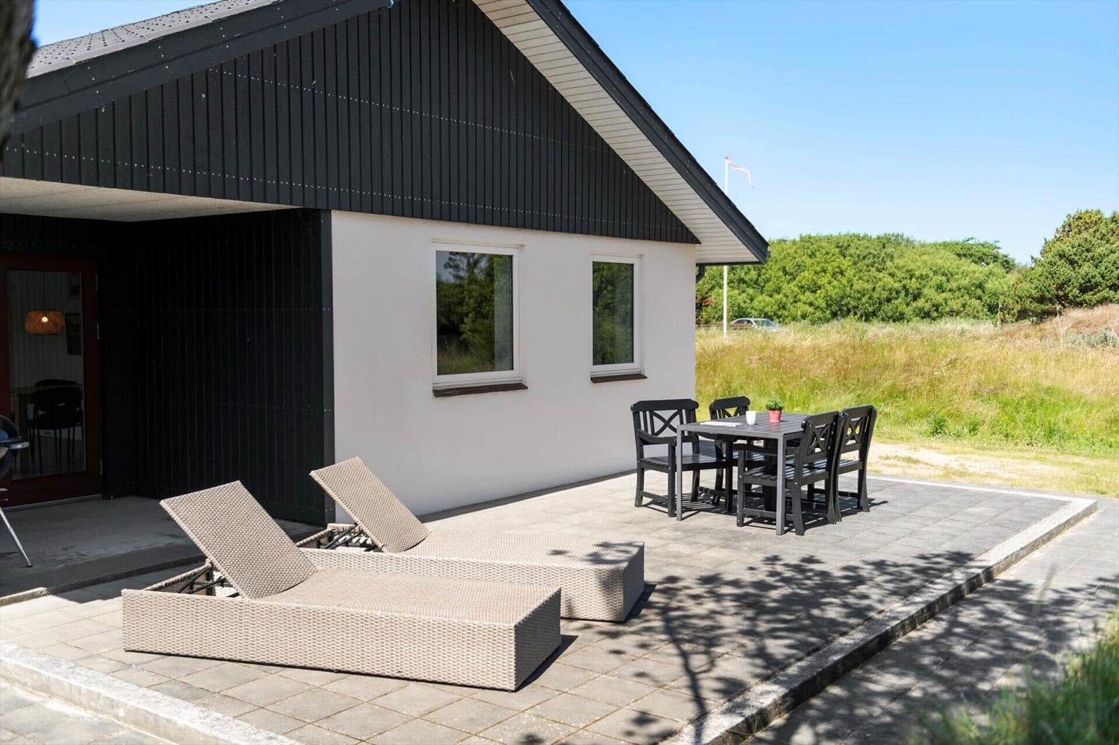 Terrasse mit Liegen und Tisch. Haus mit schwarzem Dach und weißen Wänden.