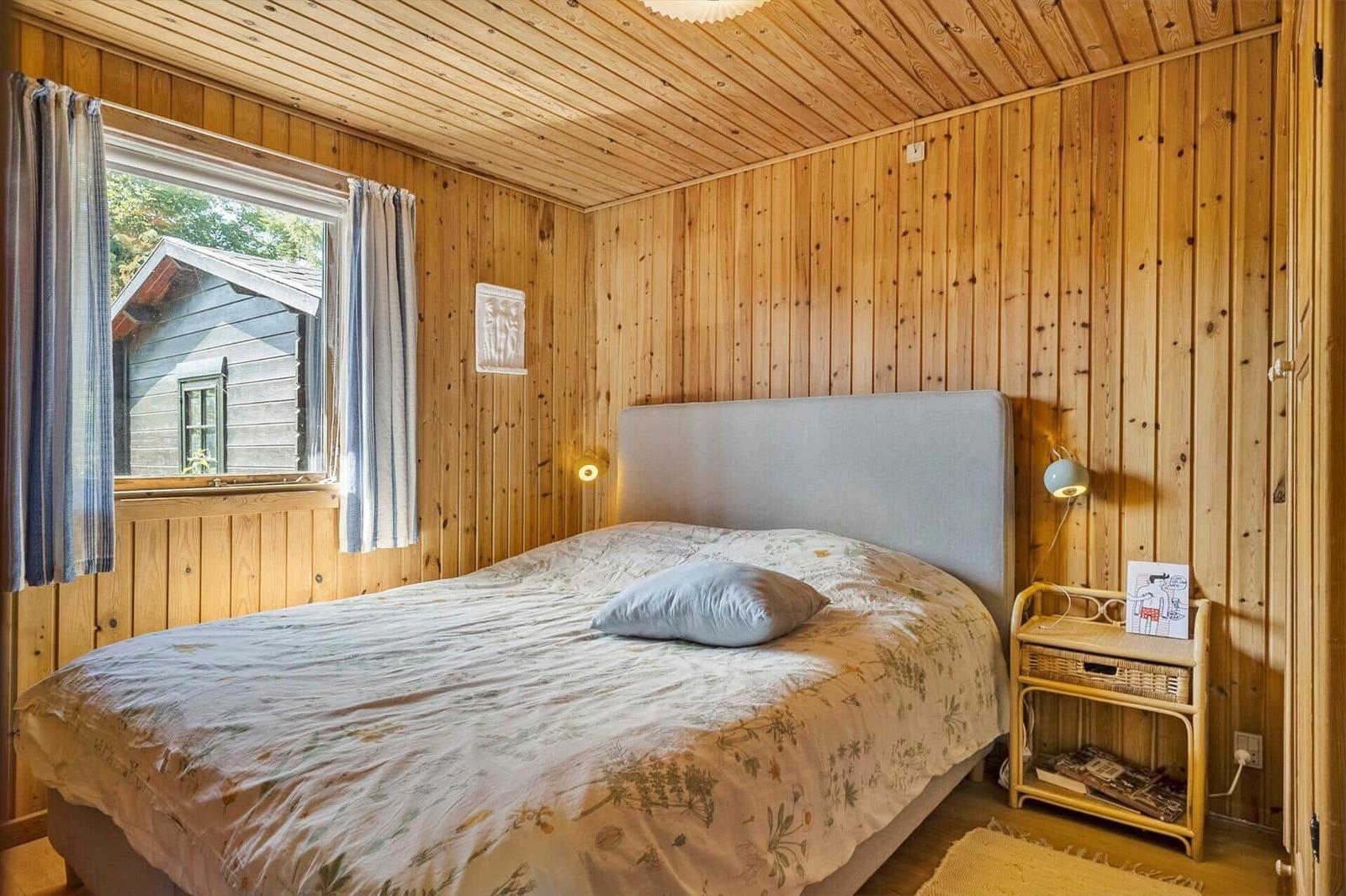 Schlafzimmer mit Holzwänden, Bett und Fensterblick auf eine Hütte.