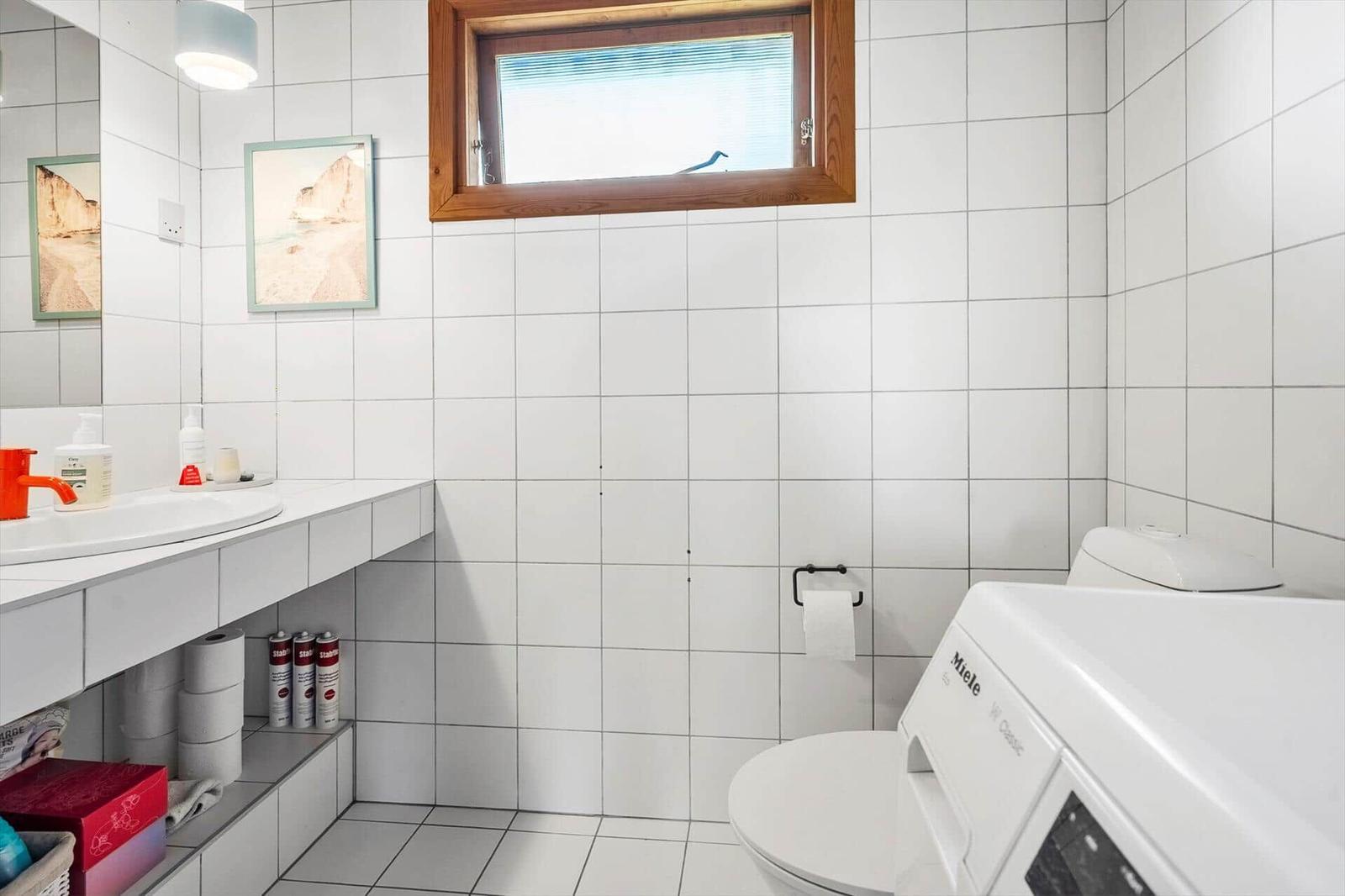 Badezimmer mit Waschbecken, Toilette und Waschmaschine. Weiße Fliesen und Holzfenster.