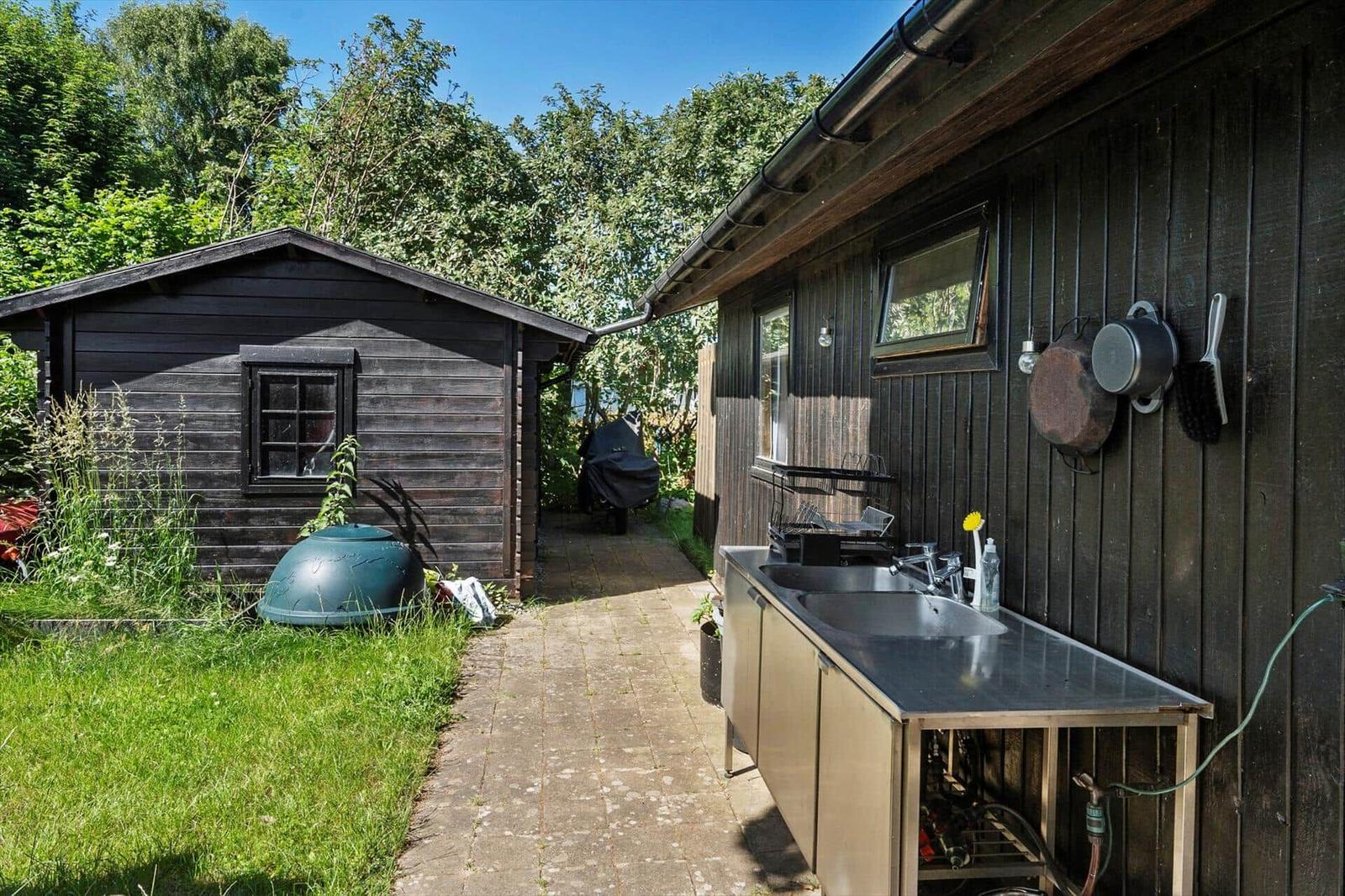 Außenküche mit Spüle und Hängeware an Holzhauswand. Neben einem Gartenhaus mit Gras und Pflanzen.