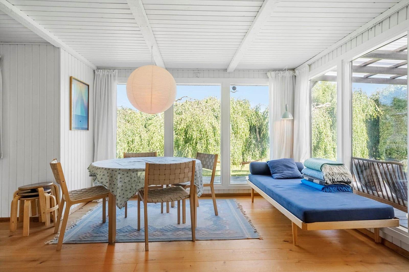 Ein Raum mit Tisch, Stühlen und Liegeplatz. Große Fenster zeigen Garten.