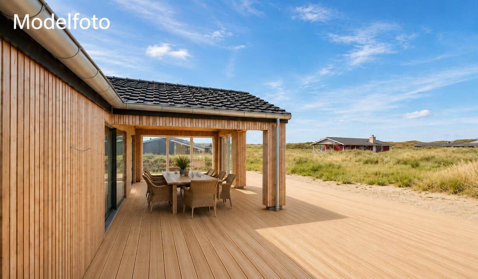 Holzterrasse mit Tisch und Stühlen unter Dach. Blick auf Dünen und Ferienhaus.