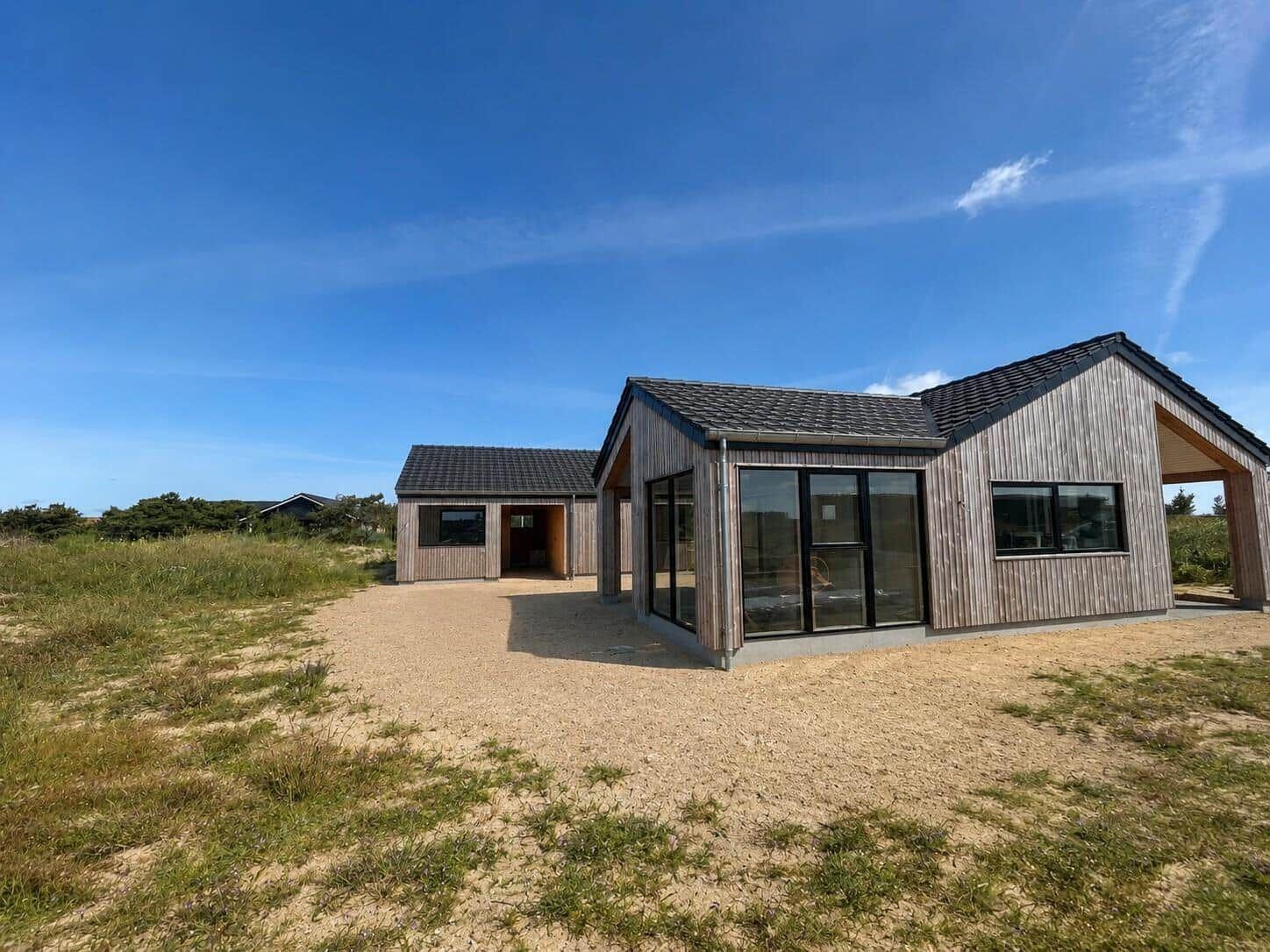 Moderne Holzhütte mit großem Fensterbereich und Dachüberstand. Grasbewachsener Boden und klarer Himmel.
