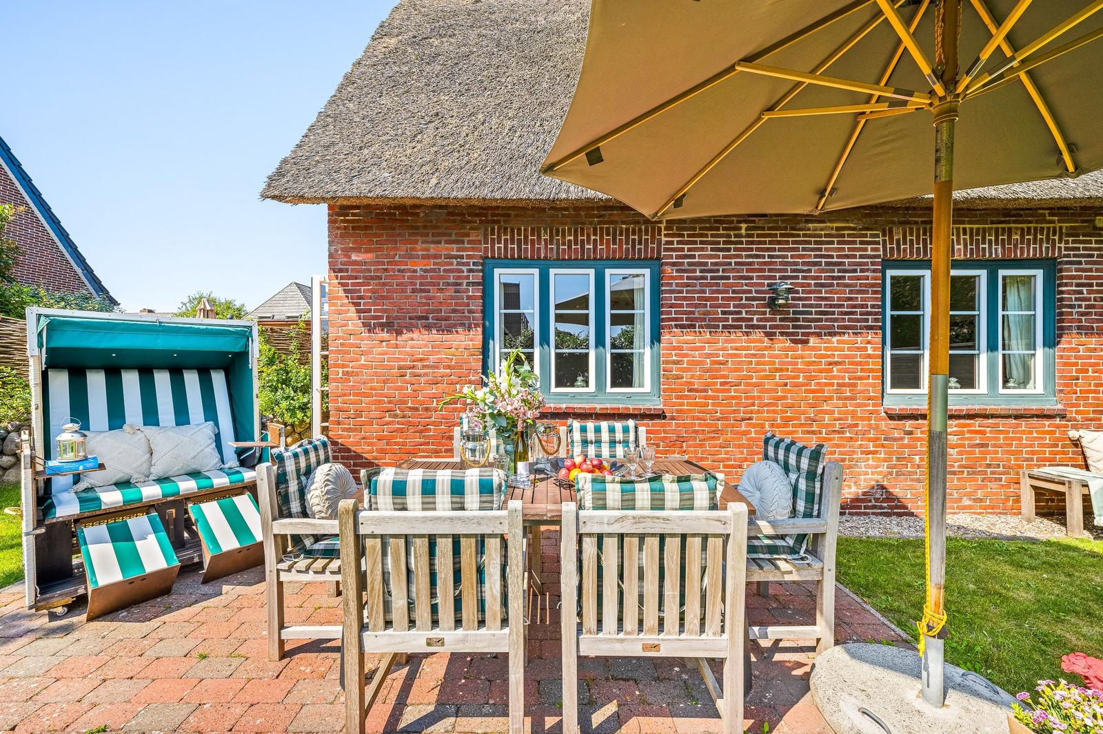 Terrace with table, chairs, and umbrella in front of a brick house with thatched roof.