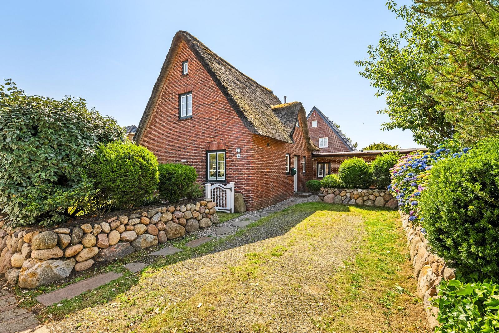 Red brick house with thatched roof, paved path, and stone walls with flower beds.