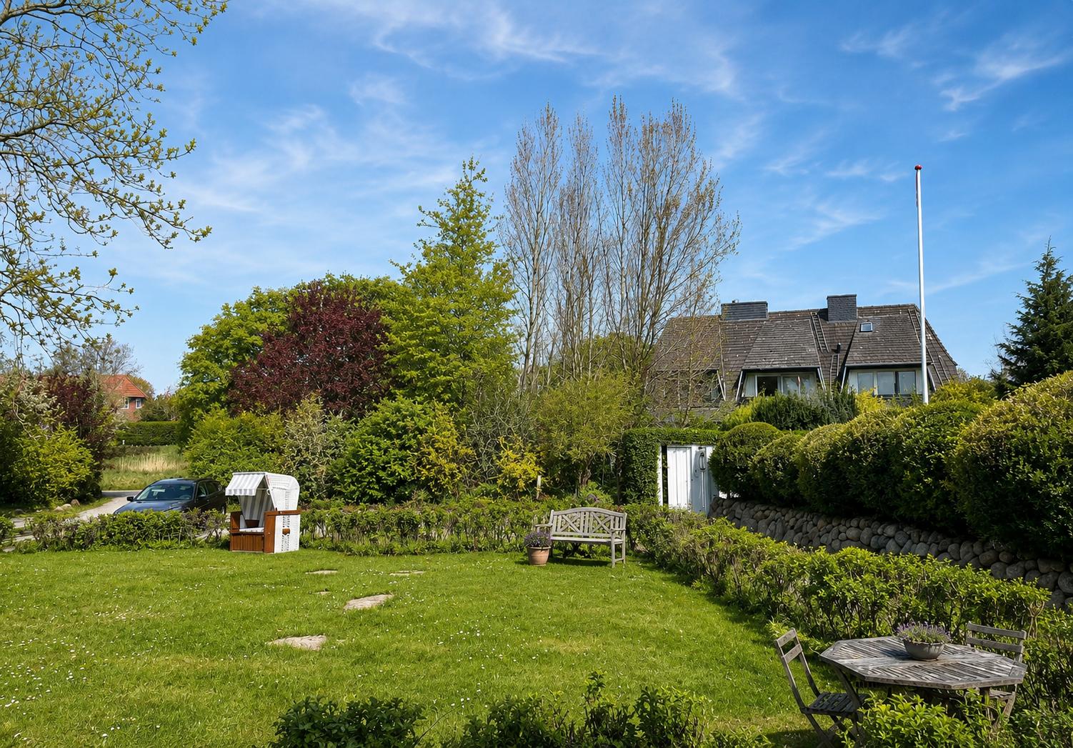 Haus mit Garten, Bäumen und einem Strandkorb. Holztisch und Stühle im Vordergrund.