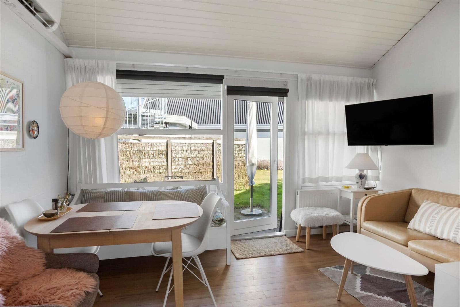 Wohn- und Essbereich mit Sofa, Tisch und Fernseher. Durchgang zur Terrasse mit Sonnenschirm.