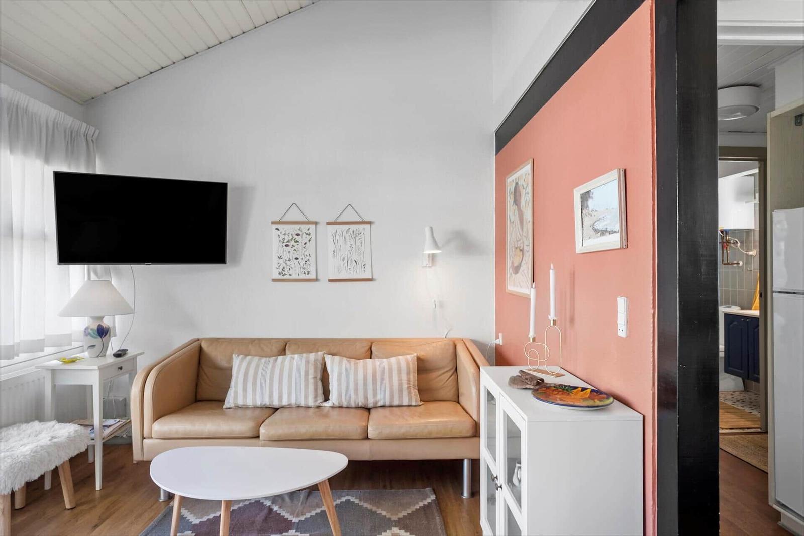 Wohnzimmer mit Sofa, Fernseher und Tisch. Wand in Rosa. Küche im Hintergrund.