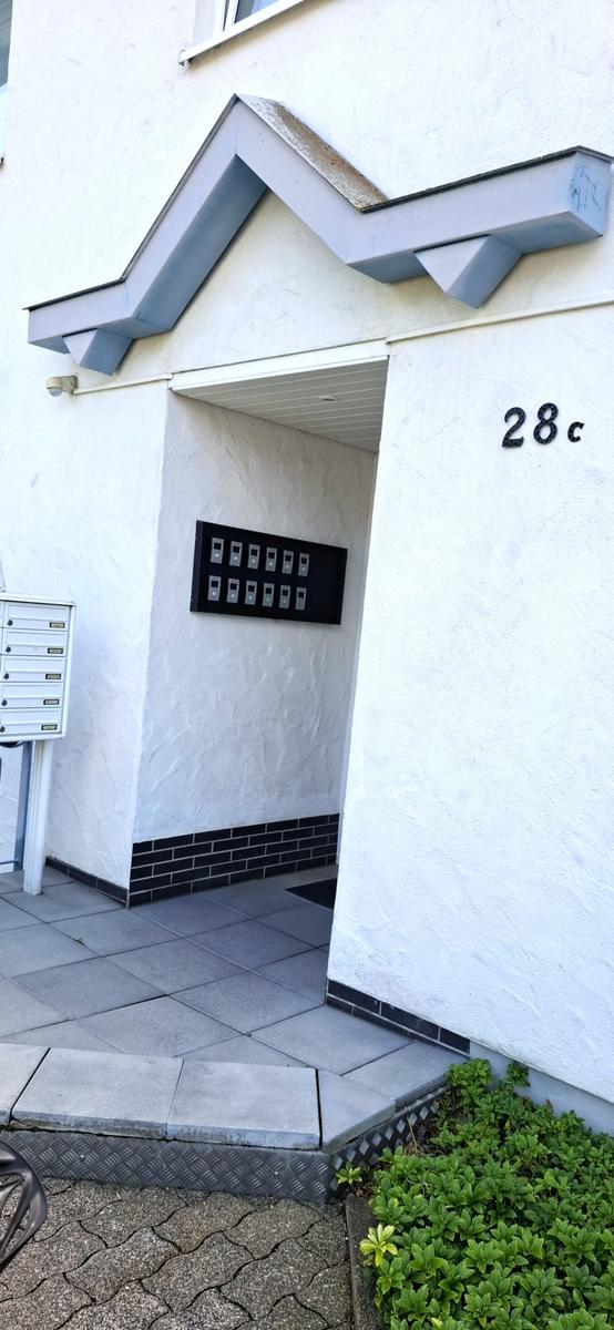 Eingangsbereich mit Hausnummer 28c und Briefkasten. Weiße Wand, graue Fliesen und Pflanzen.