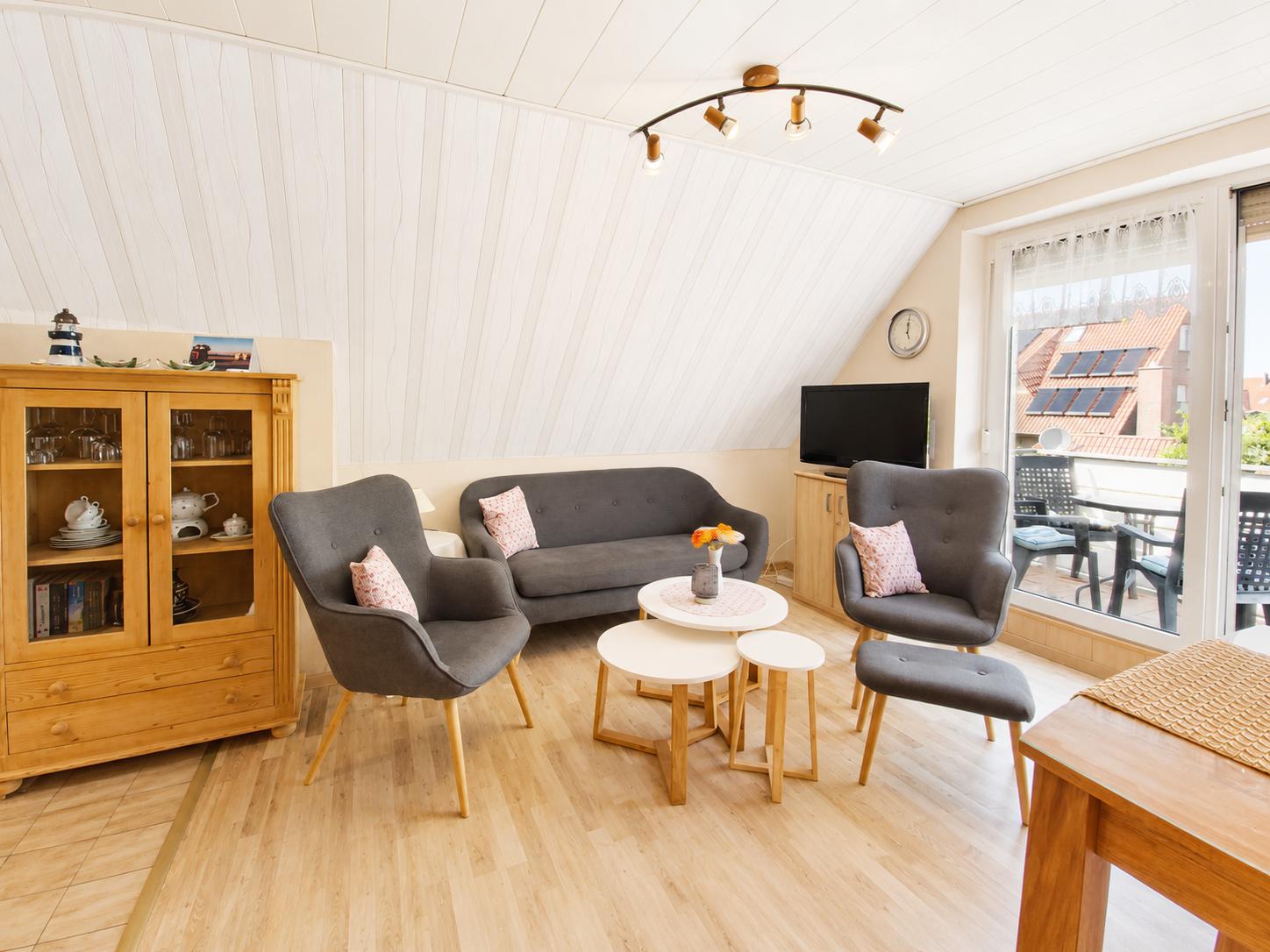 Wohnzimmer mit grauer Couch, zwei Sesseln und Holzmöbeln. Balkon mit Blick auf Dächer.