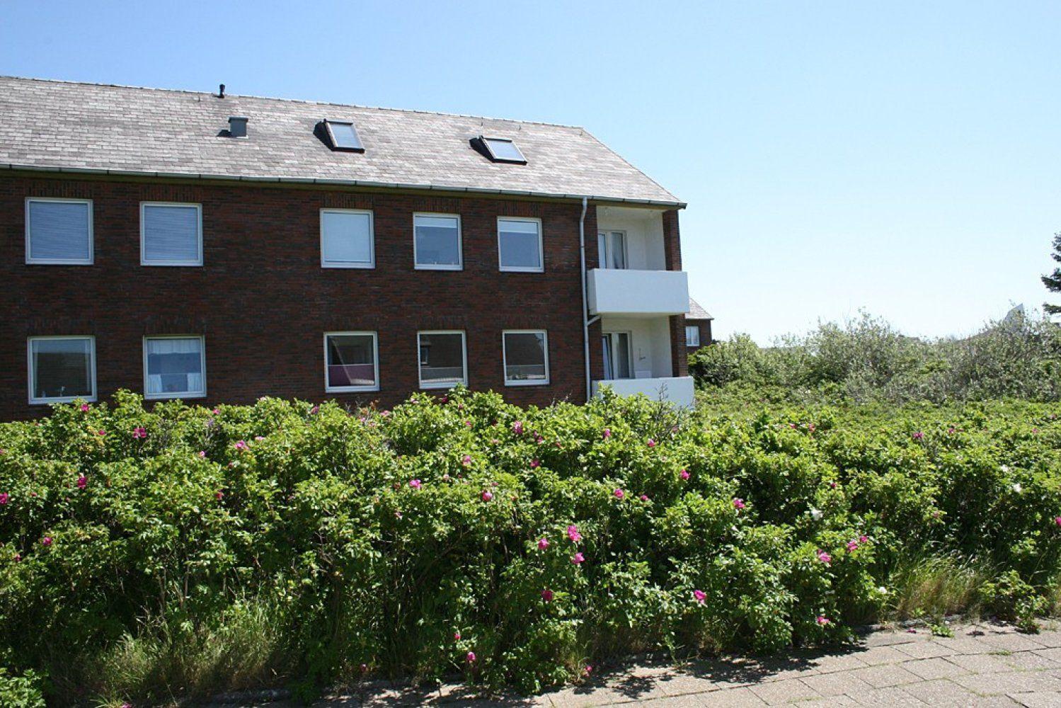 Two-story building with red brick and balcony. Front has a rose hedge.
