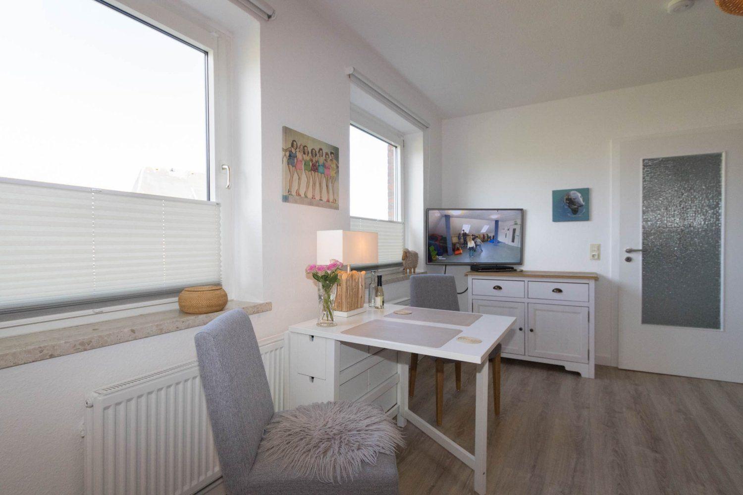 A room with a table, chairs, TV, and windows. Wooden floor and white furniture.