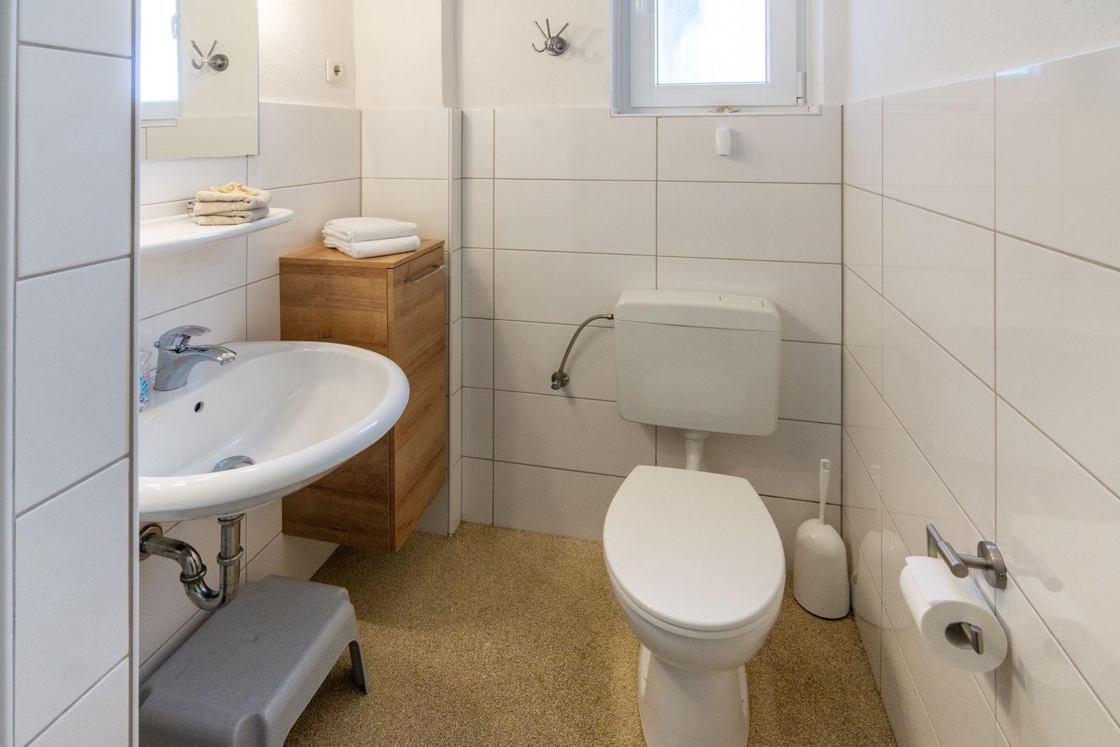 Badezimmer mit Toilette, Waschbecken und Spiegel. Holzschrank und Toilettenbürste sichtbar.
