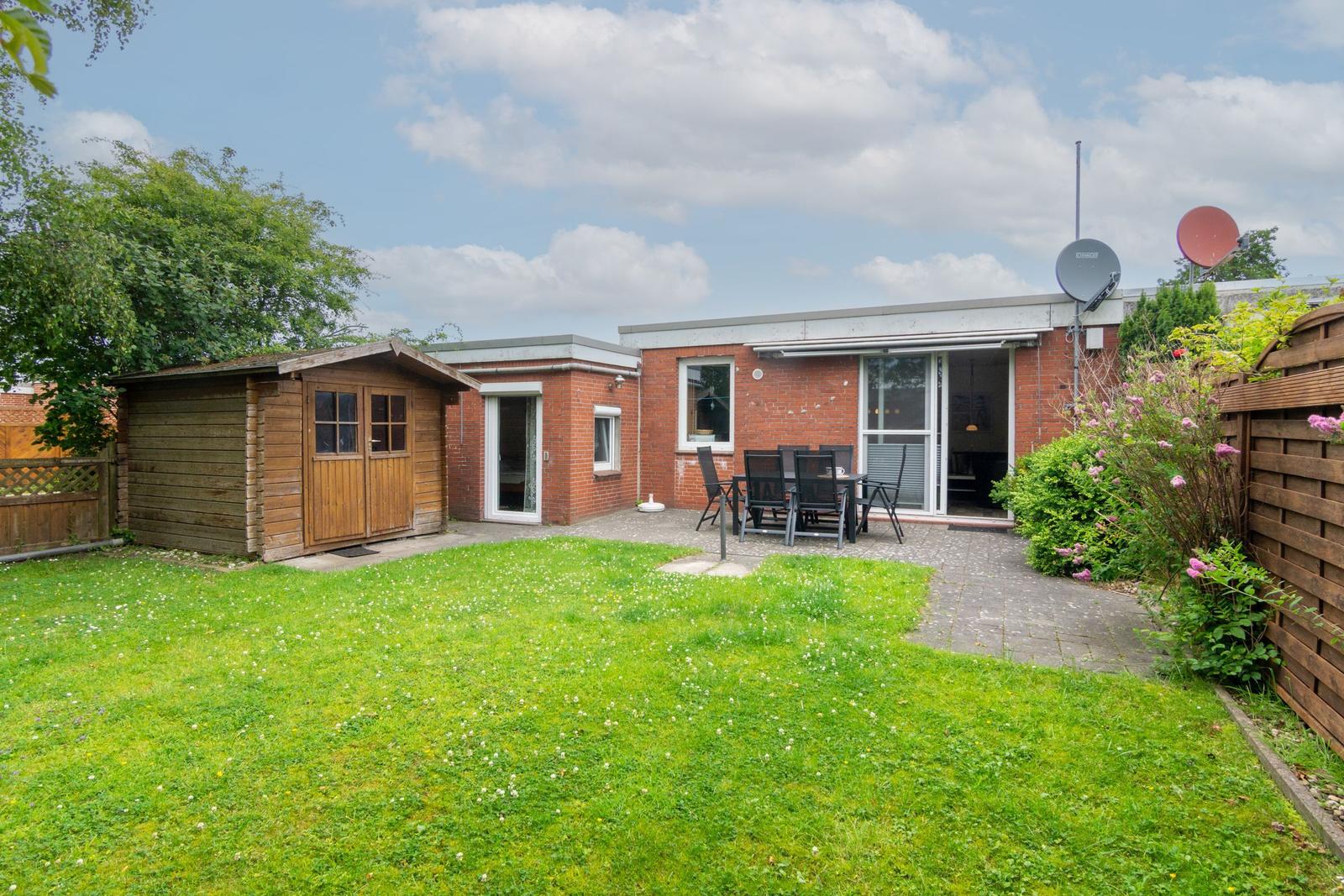 Haus mit Garten, Terrasse und Holzschuppen. Satellitenschüssel und Blumenbeete sichtbar.