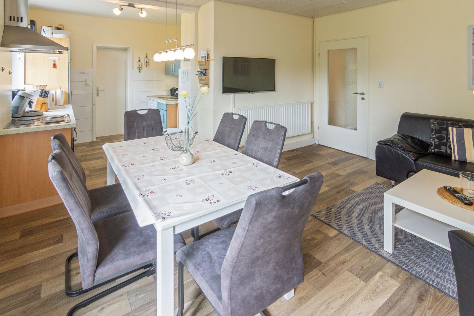 Essbereich mit Tisch und Stühlen, Wohnbereich mit Sofa und Fernseher, Küche im Hintergrund.