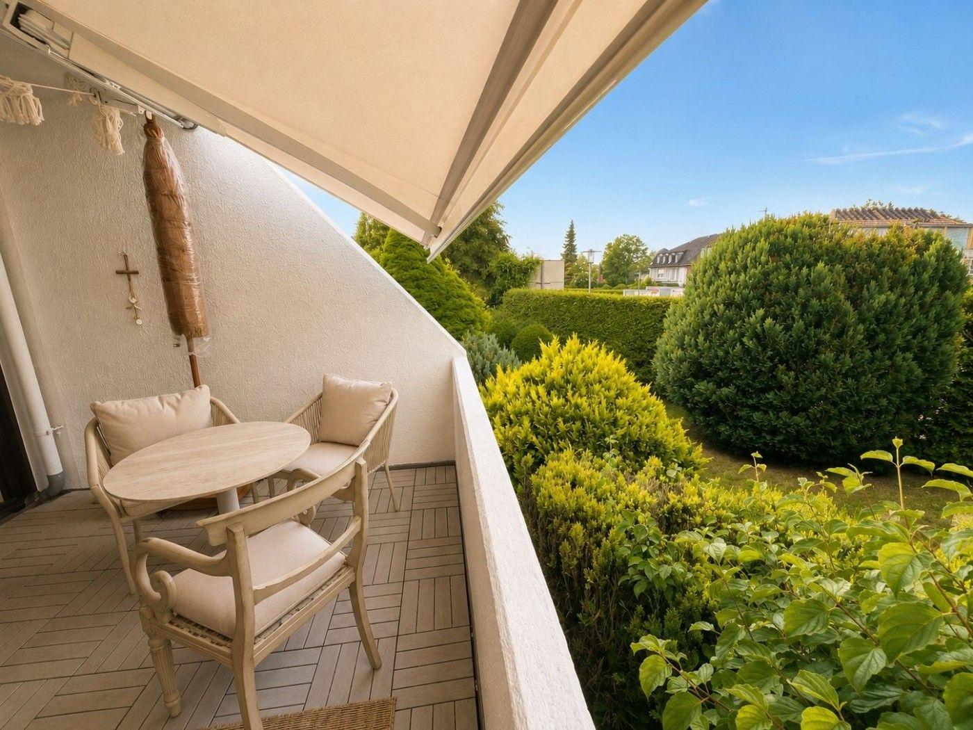 Terrasse mit Tisch, Stühlen und Markise. Blick auf grüne Hecken und einen klaren Himmel.