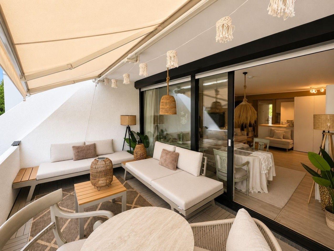 Terrasse mit Sitzgelegenheiten und Blick ins Haus. Glaswände und Hängelampen sichtbar.
