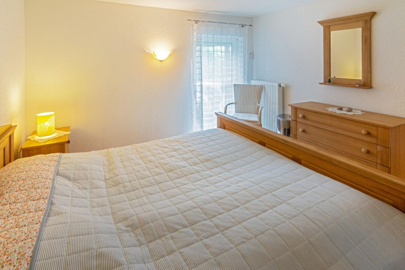 Schlafzimmer mit Holzbett, Nachttisch, Spiegel und Stuhl vor Fenster.