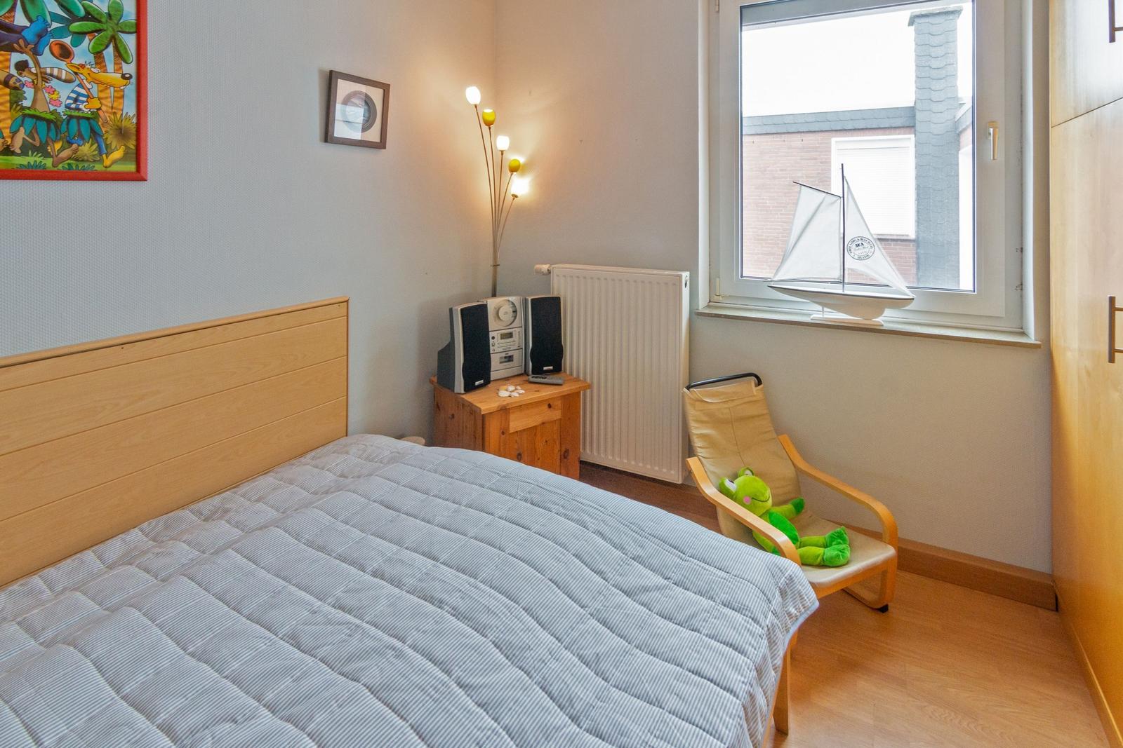 Schlafzimmer mit Bett, Nachttisch, Stuhl und Fenster. An der Wand hängt ein Bild.