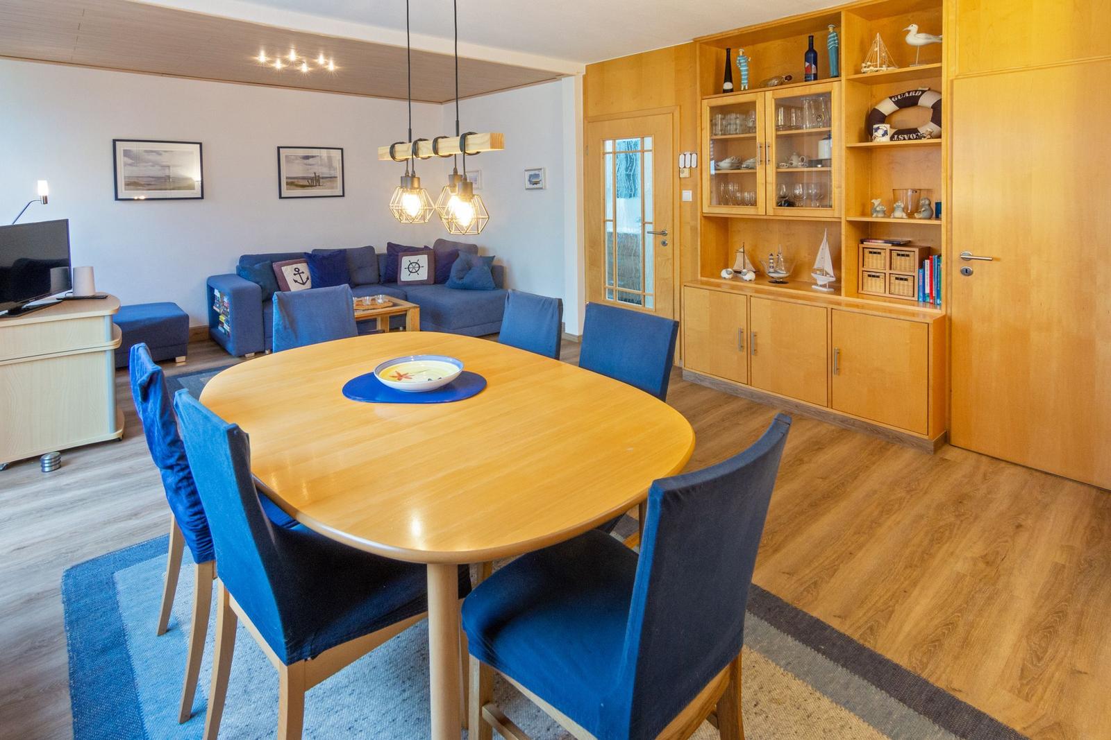 Essbereich mit runden Tisch und blauen Stühlen. Hintergrund: Wohnbereich mit Sofa und Holzmöbel.