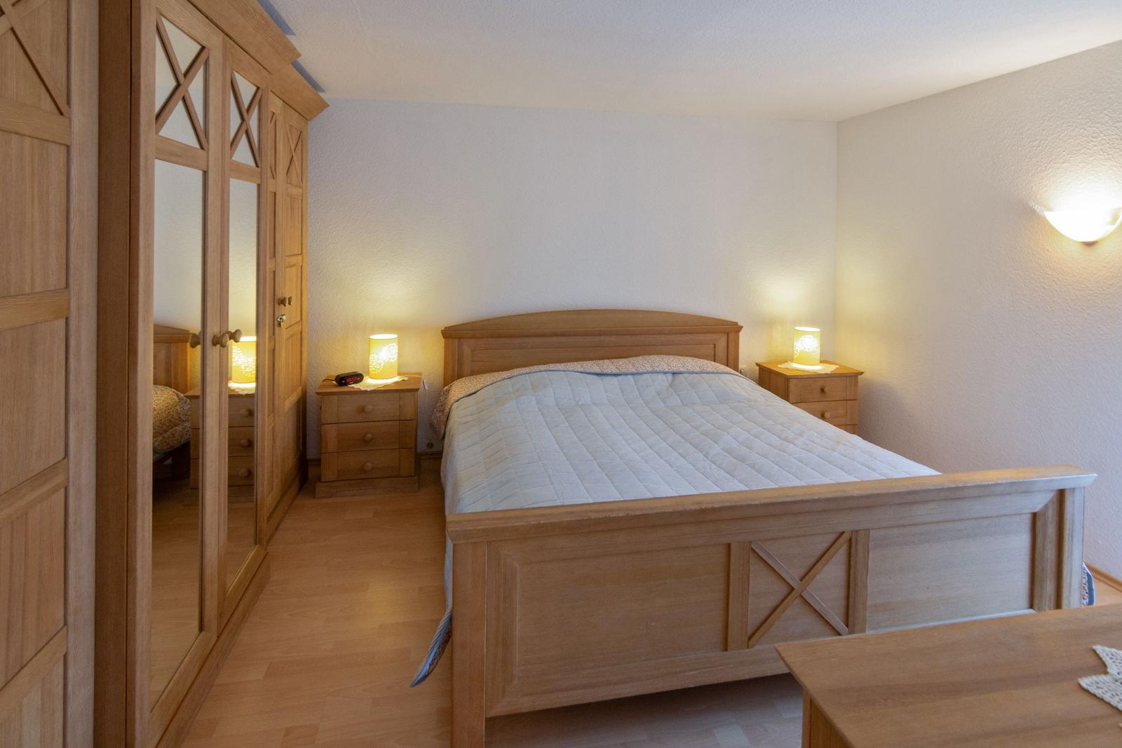 Schlafzimmer mit Holzbett, Nachttischen und Kleiderschrank. Beleuchtung durch Wandlampe und Lampen.