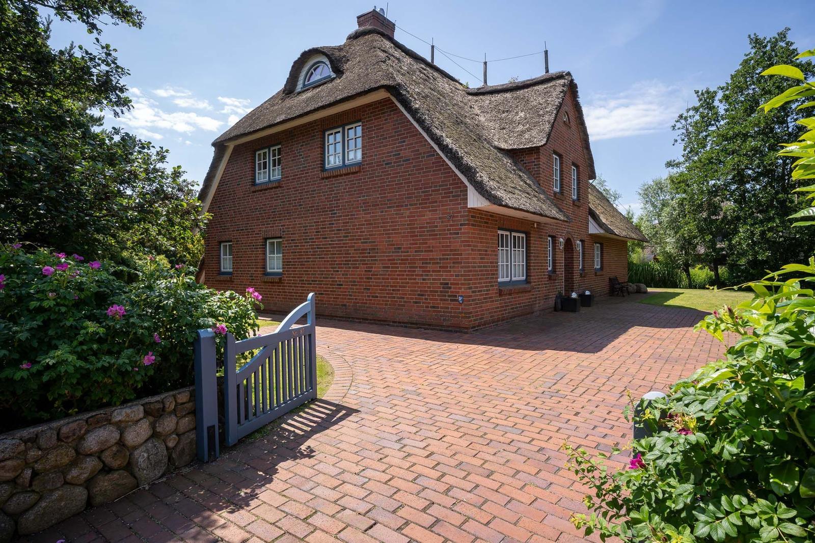 Rotes Backsteingebäude mit Strohdach und gepflastertem Hof. Umgeben von Grünflächen und Blumen.