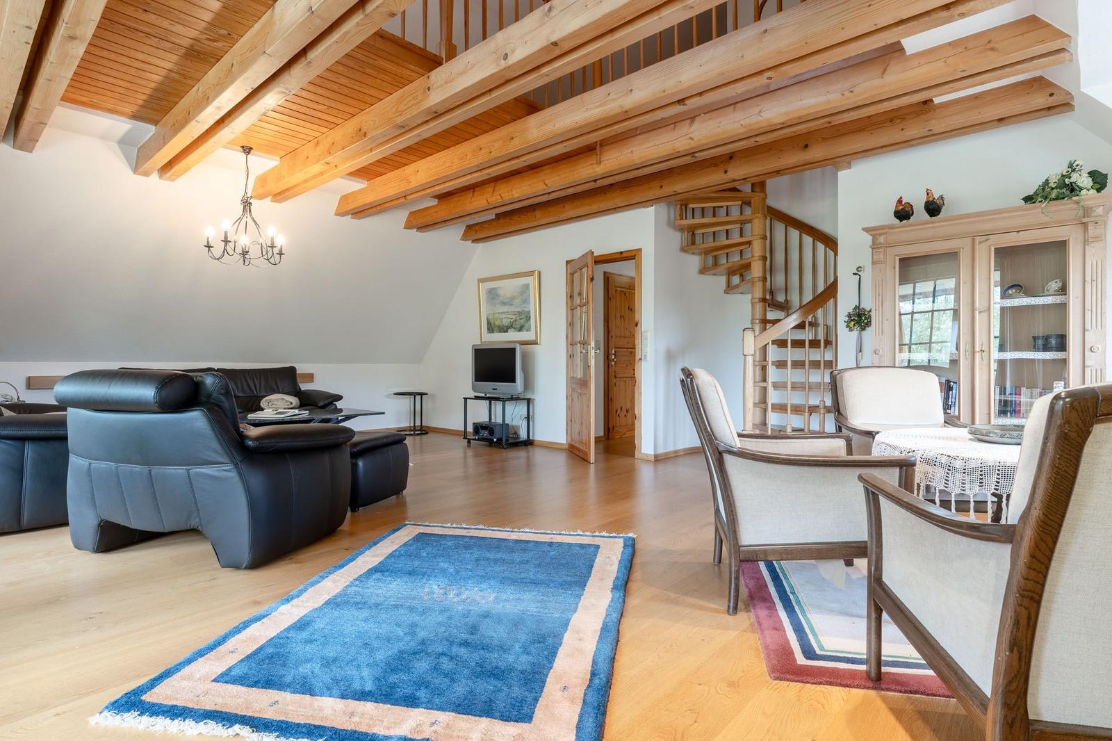 Wohnzimmer mit Holzbalken, Sofa, Stühlen und Teppich. Treppe führt zur oberen Etage.