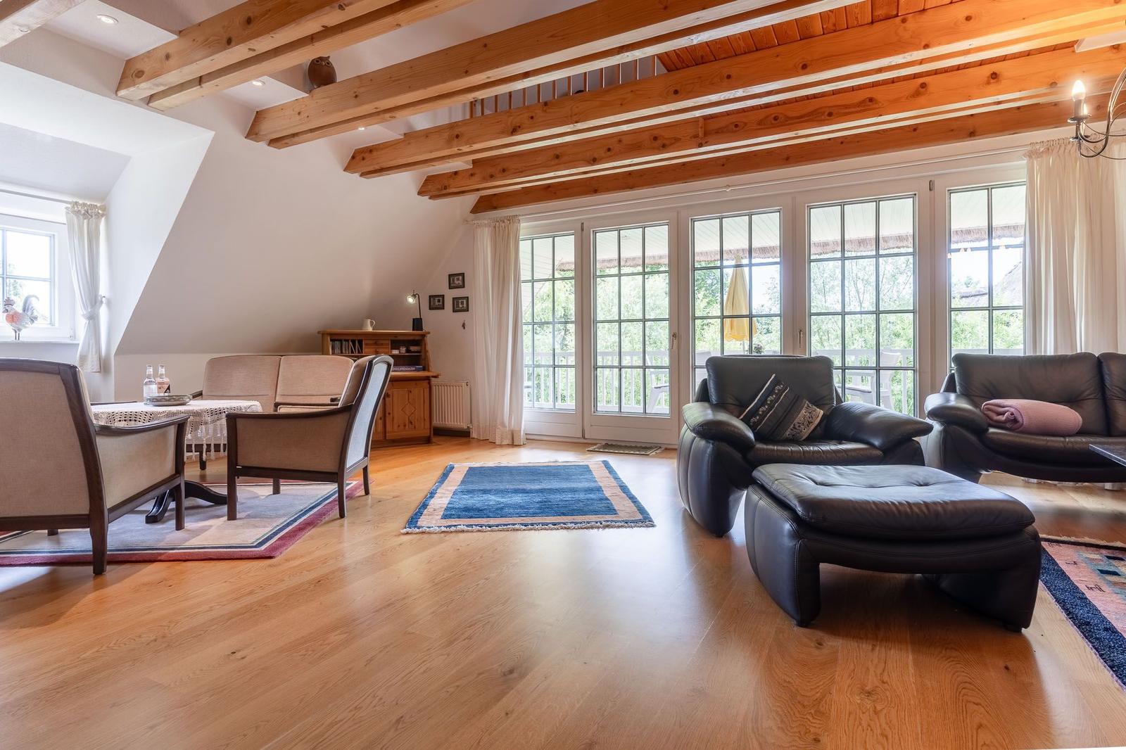 Wohnzimmer mit Holzbalken, großen Fenstern und Ledersofas auf Holzboden.
