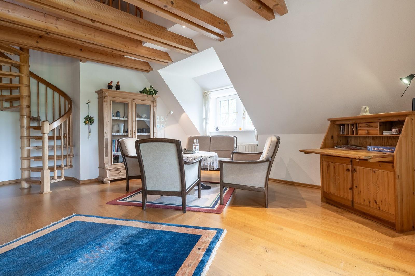 Wohnraum mit Holzdecke, Spiraltreppe und Holzmöbeln. Zwei Teppiche und Sitzgruppe sind sichtbar.