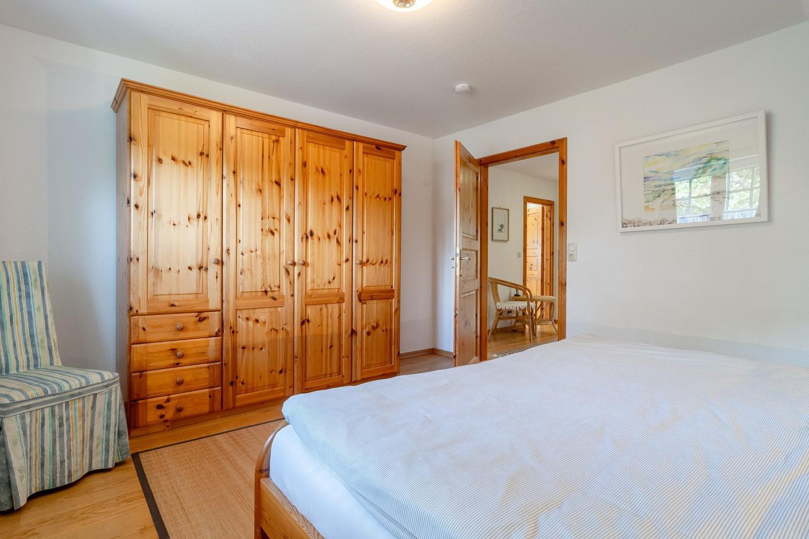 Schlafzimmer mit Holzschrank, Bett und Sessel. Tür führt in angrenzendes Zimmer.