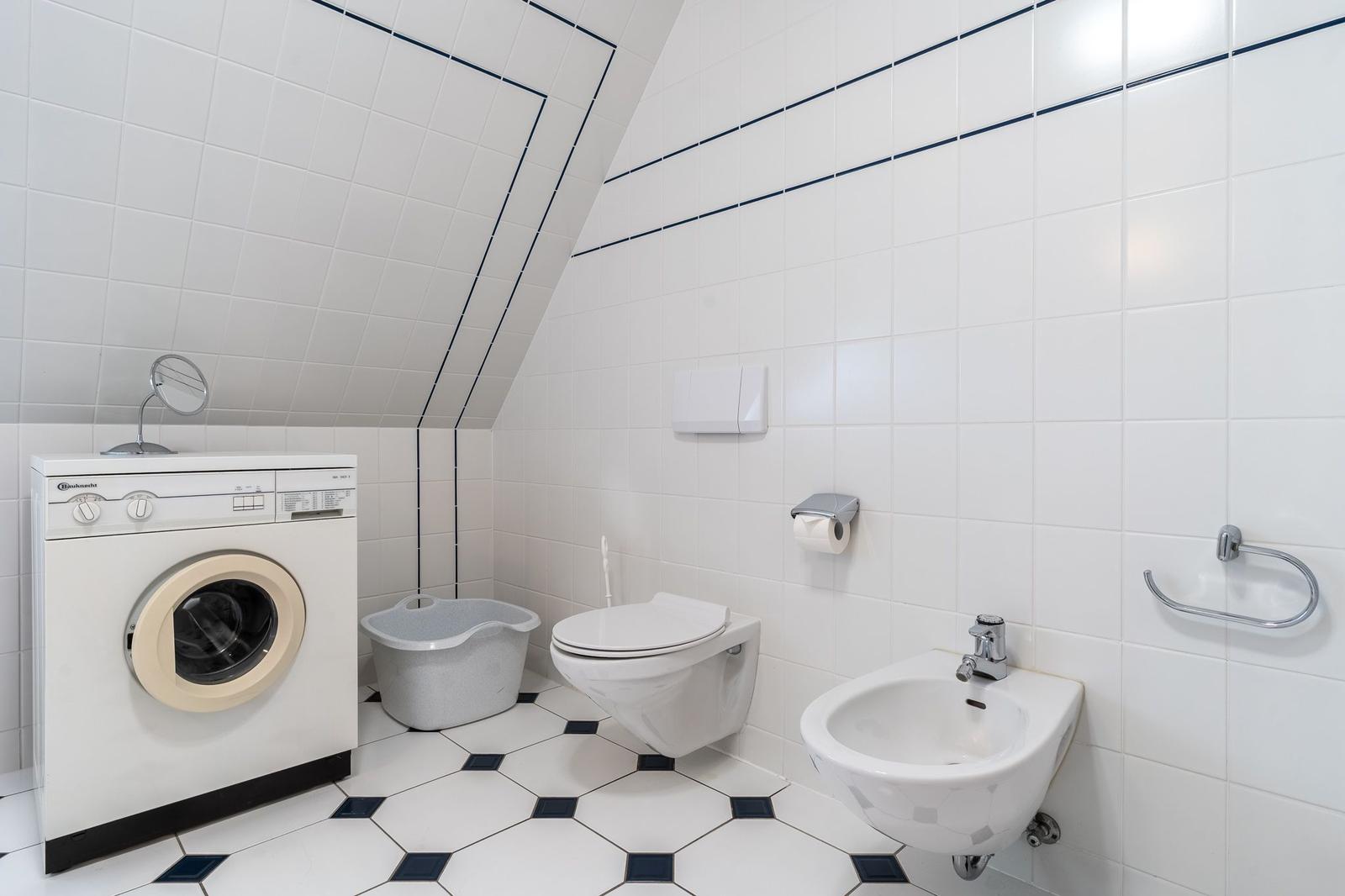 Badezimmer mit Waschmaschine, Toilette und Bidet. Weiße Fliesen mit dunklen Akzenten.
