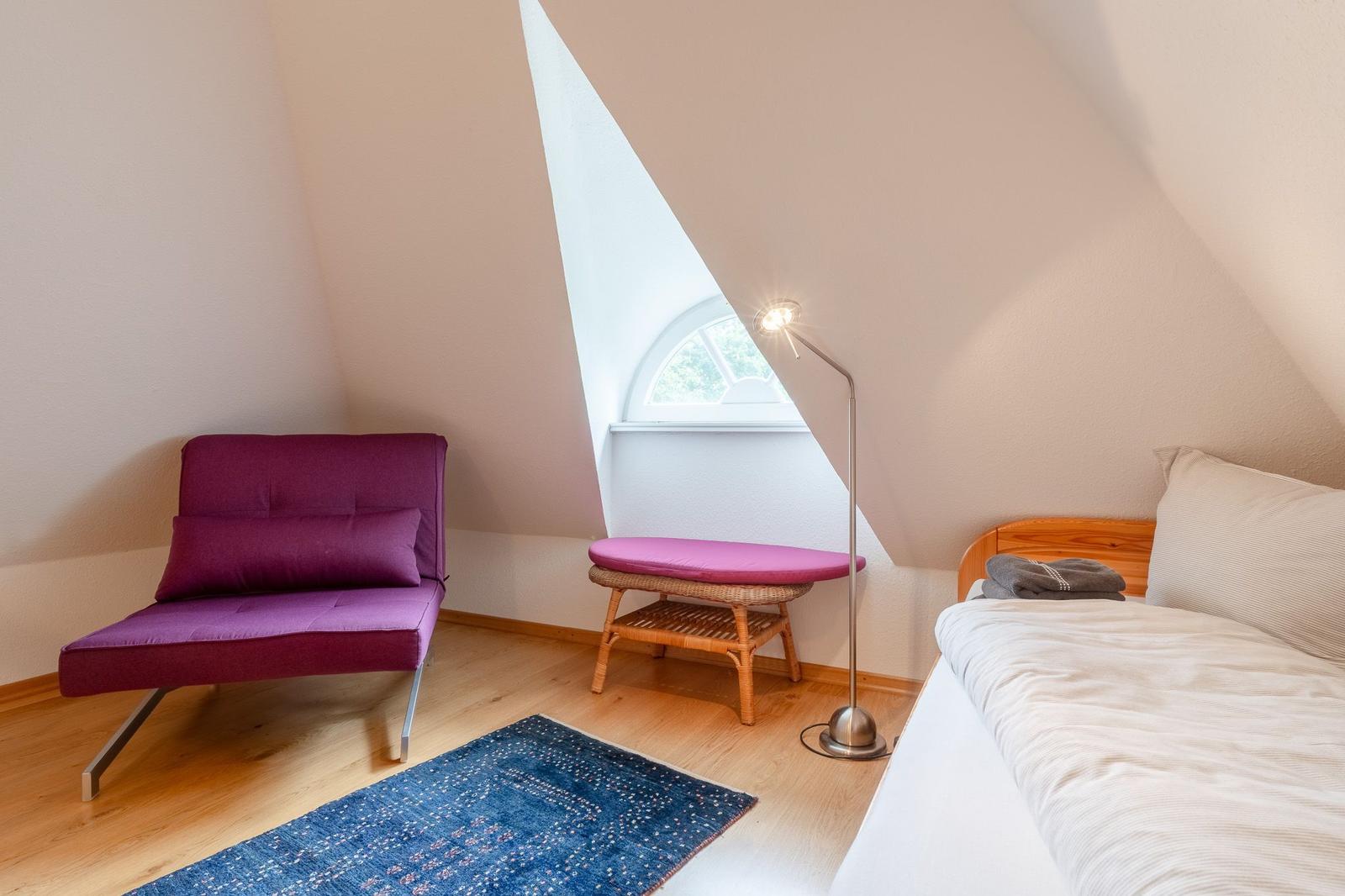 Schlafzimmer mit Bett, violettem Sessel und Fenster unter Dachschräge.