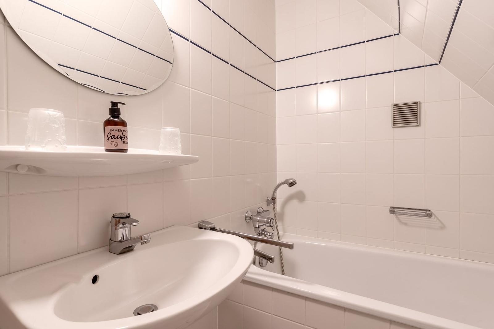 Badezimmer mit Badewanne, Waschbecken und runden Spiegel. Weiße Fliesen mit blauen Akzenten.