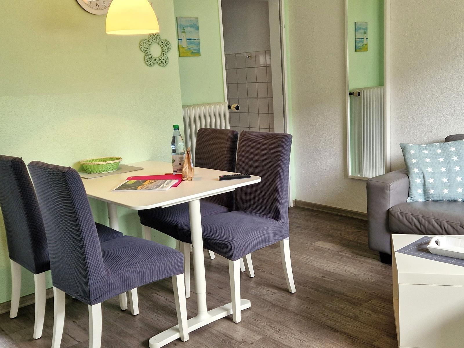 Essbereich mit Tisch und Stühlen, Sofa mit Kissen, Wandbild und Heizkörper sichtbar.