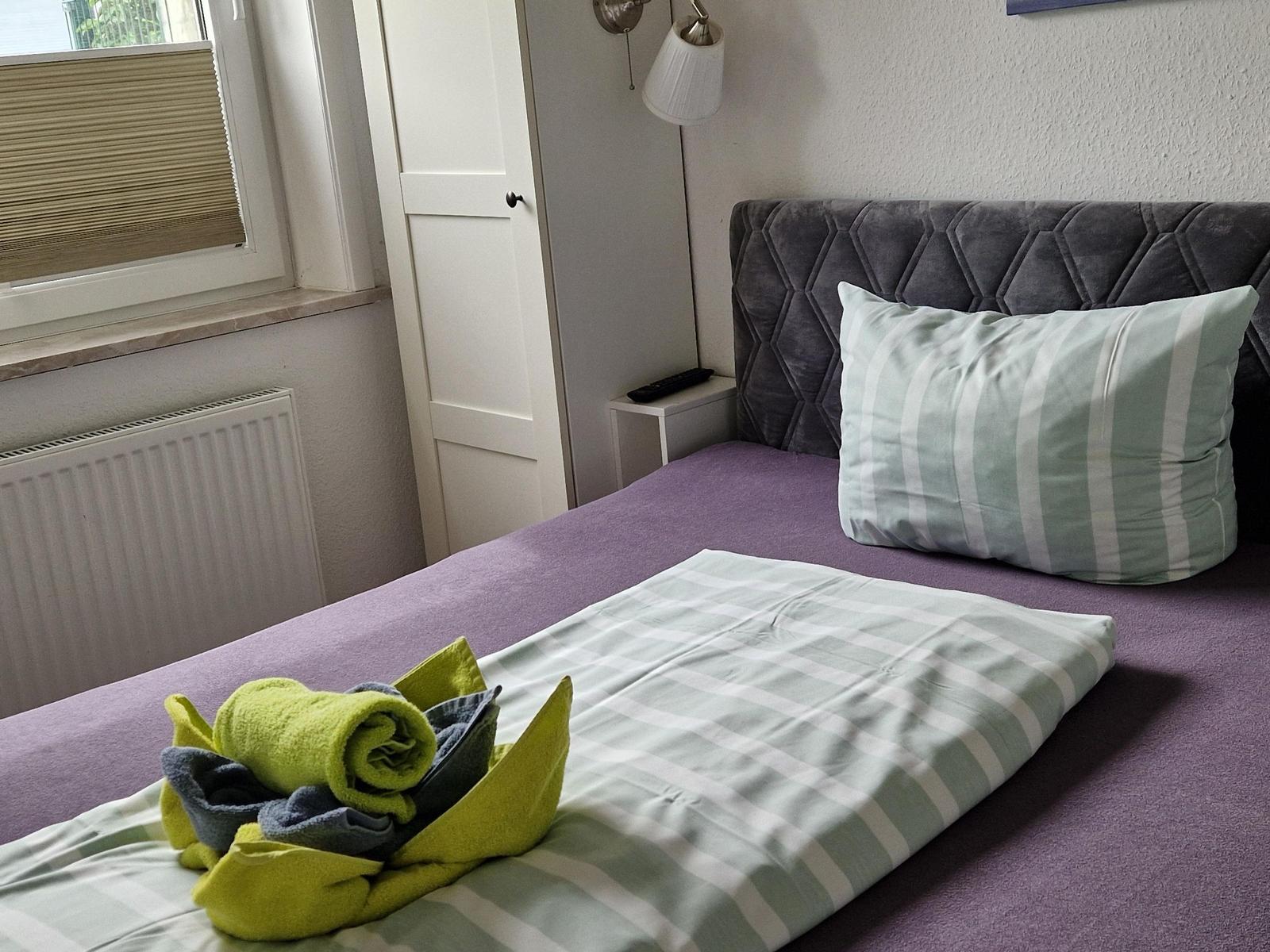 Schlafzimmer mit Bett, Kissen und Handtüchern. Fenster, Schrank und Wandlampe sichtbar.